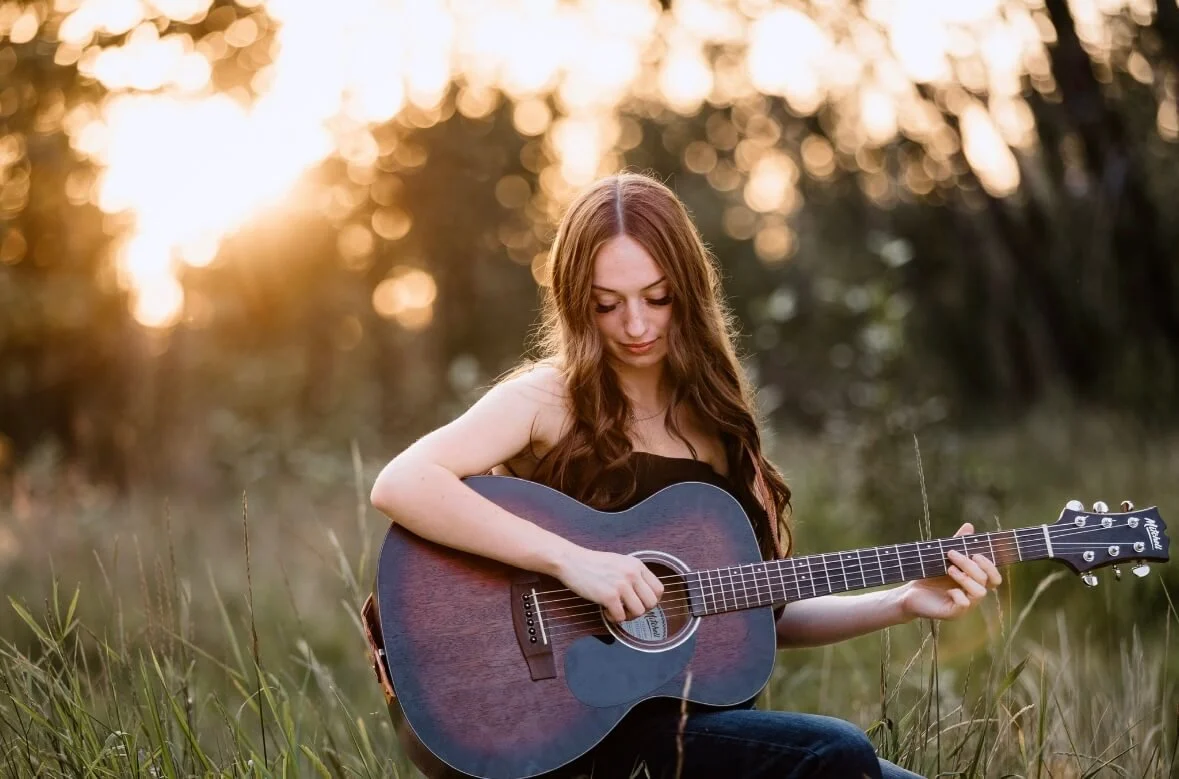 A young woman with long brown hair, wearing a black sleeveless top, sitting in a grassy field at sunset, playing an acoustic guitar.