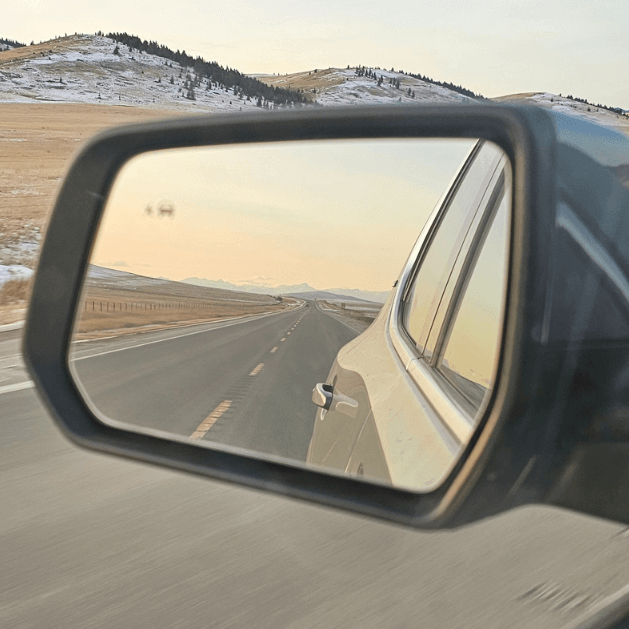 The view from a sideview mirror of a vehicle, in the mirror you can see the side of the car and the road behind, and mountains in the distance.  In front of the mirror, you can see rolling hills with some snow and trees on them.