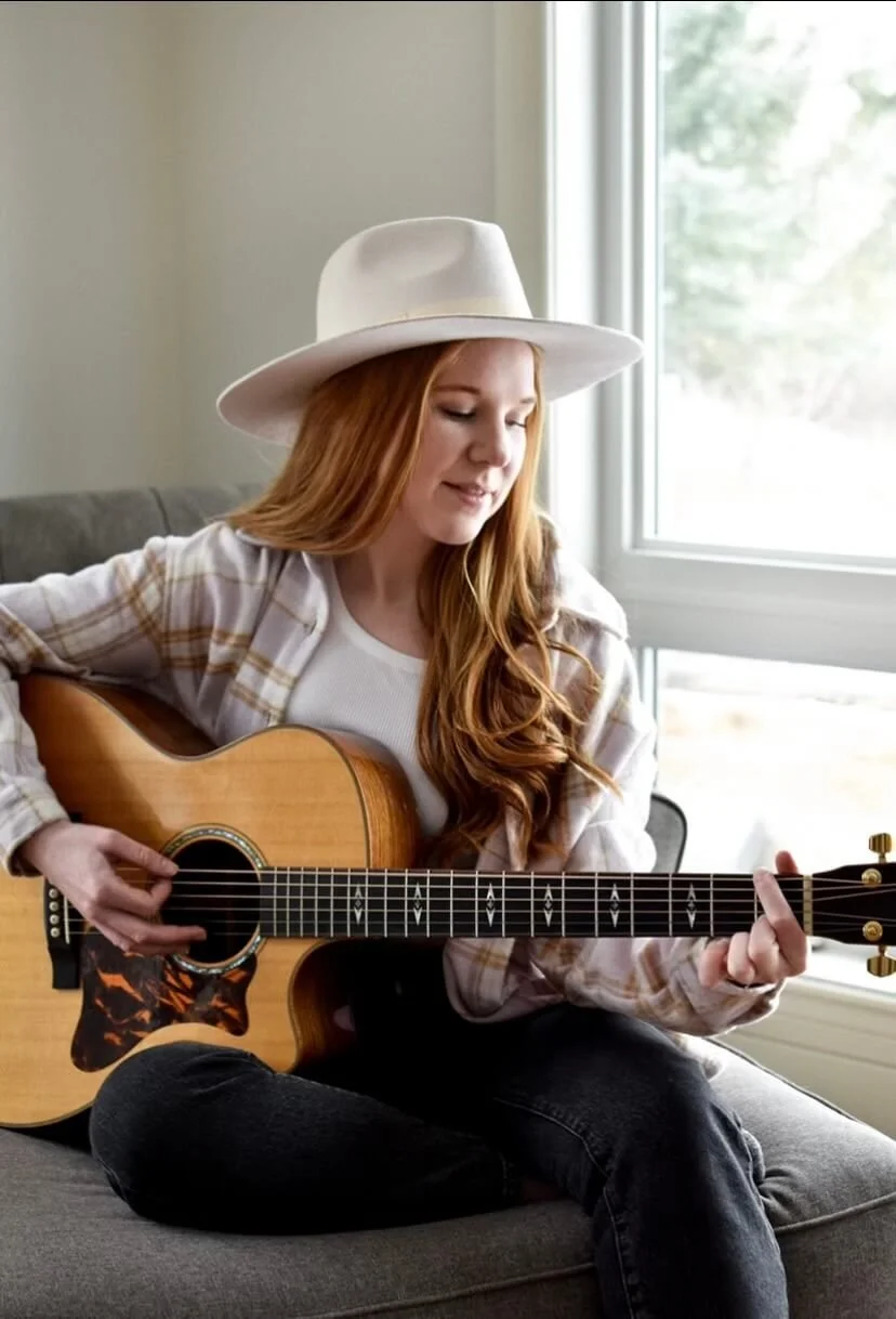 A young woman with long red hair wearing a white hat and casual plaid shirt playing an acoustic guitar while sitting on a gray couch near a window.