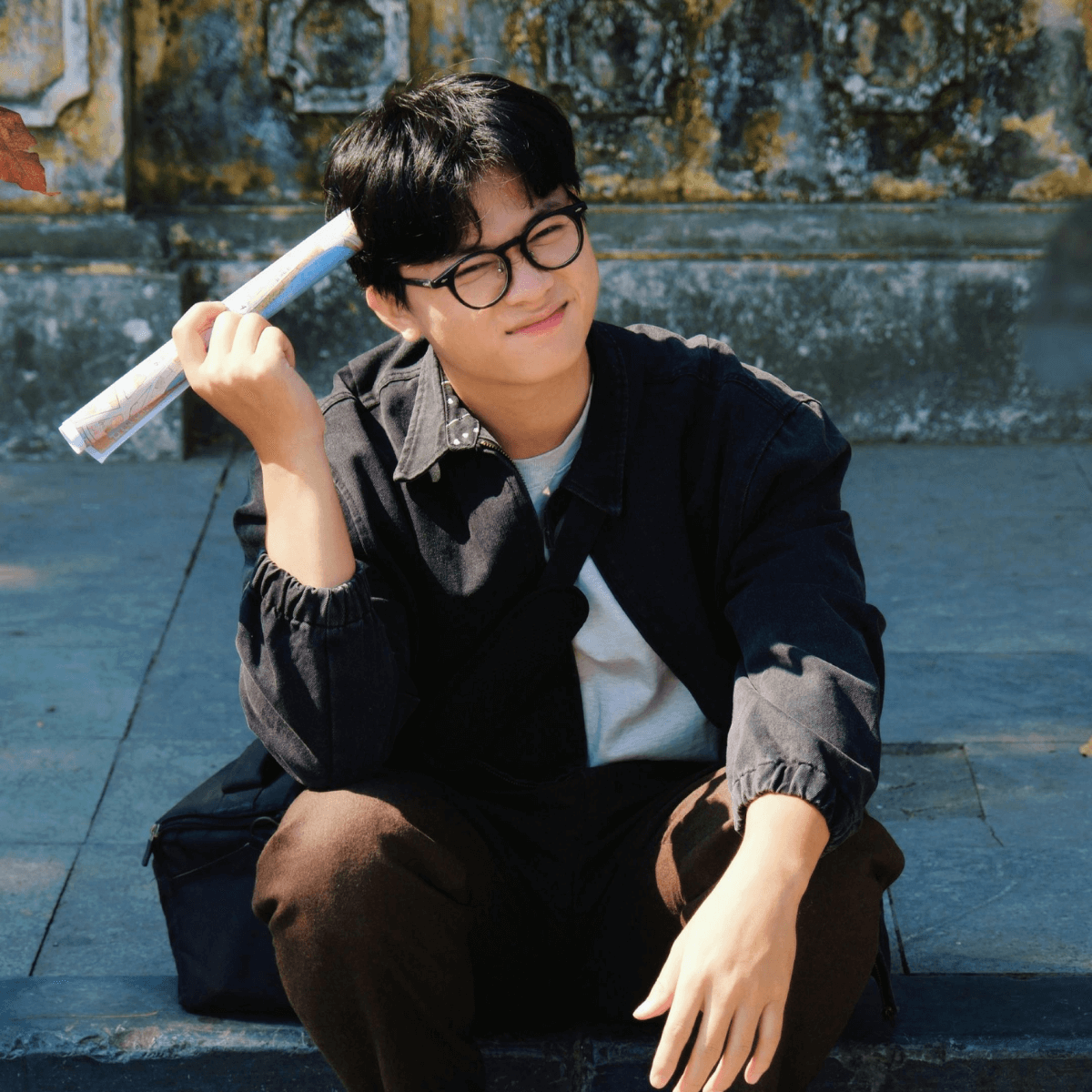 A young man sitting on the edge of a sidewalk with a rolled up piece of paper in his hand and a thoughtful look on his face.