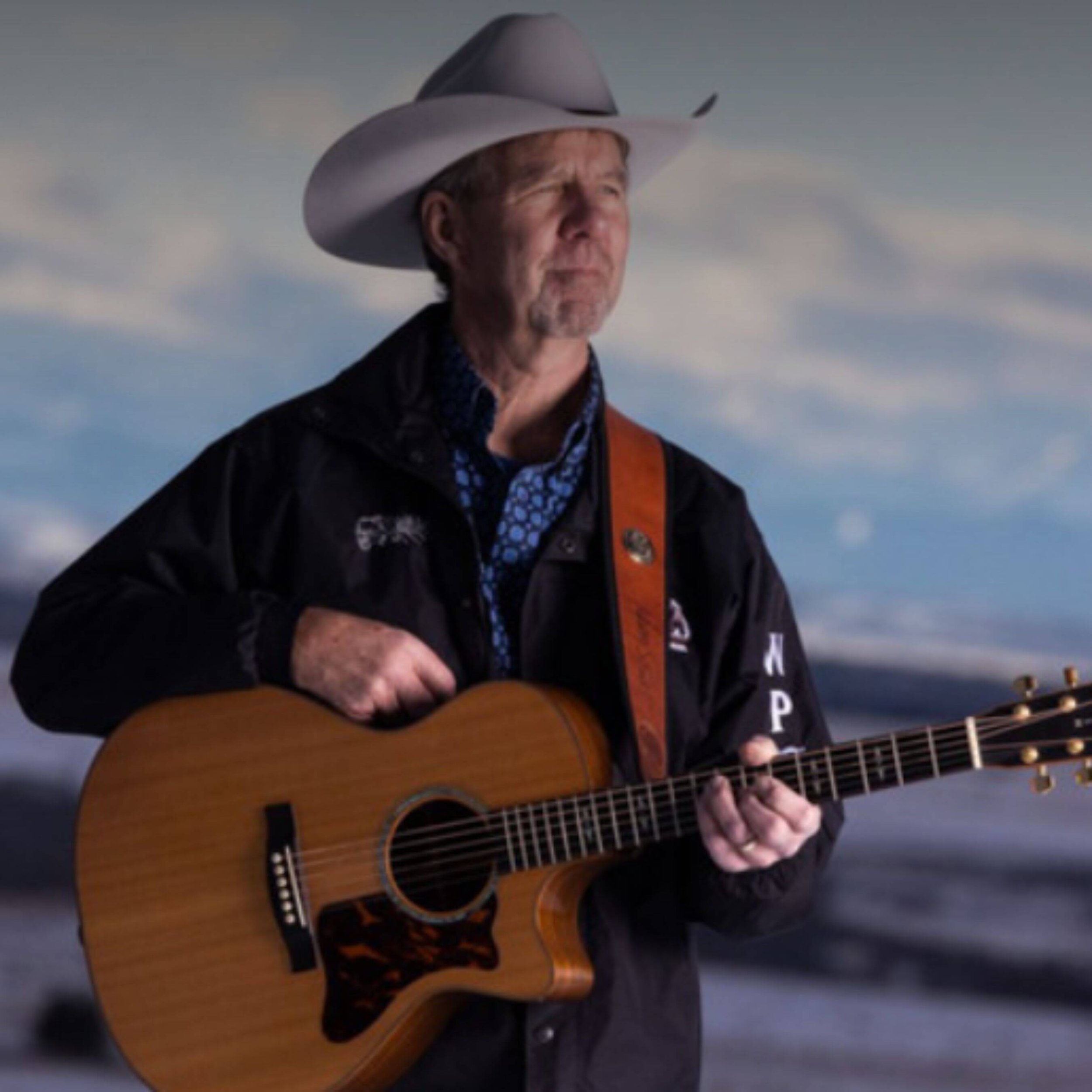 A man wearing a cowboy hat and dark jacket playing an acoustic guitar outdoors with sky and clouds in the background.