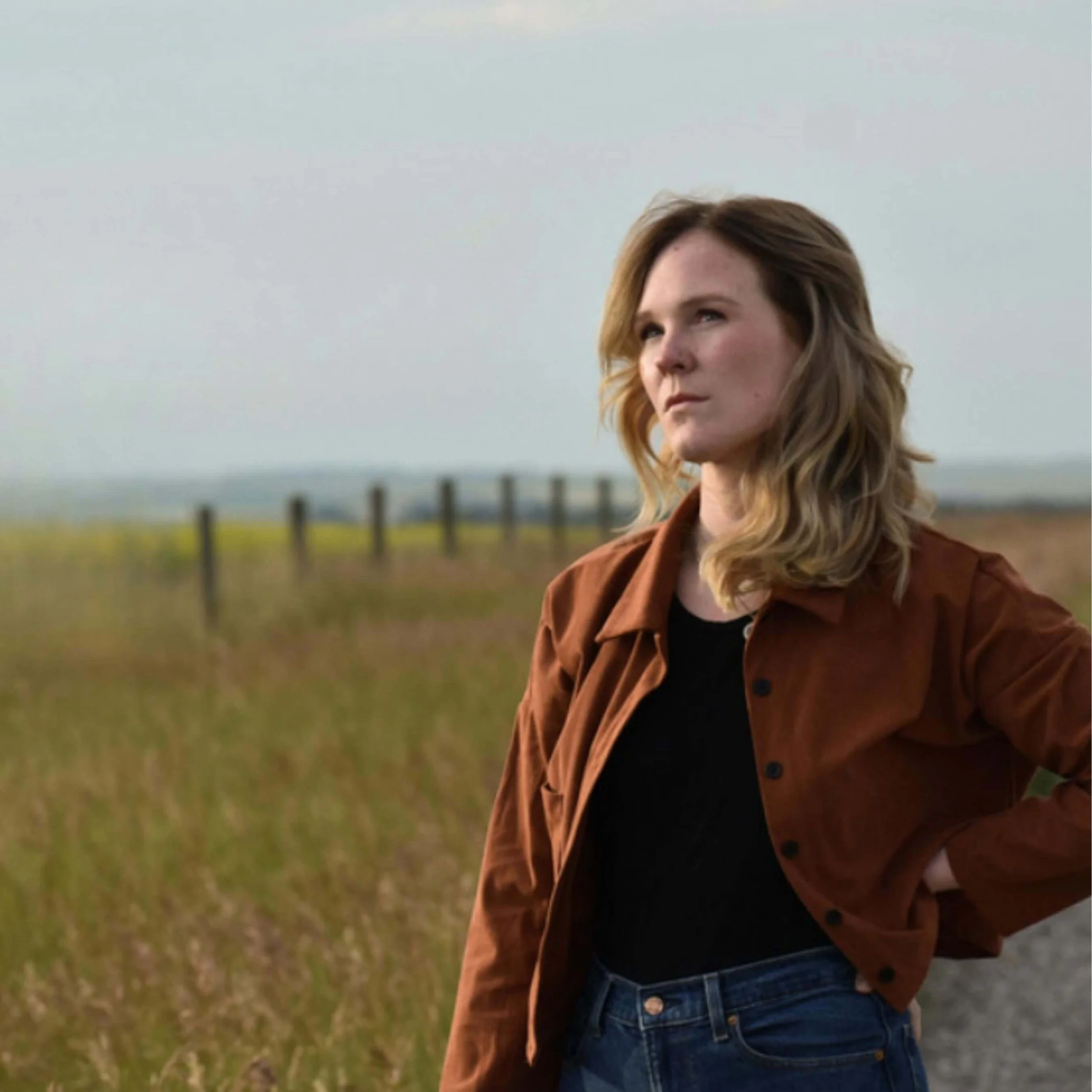 Young woman with wavy blonde hair standing outdoors in a field with a fence in the background, wearing a brown jacket over a black top and blue jeans.