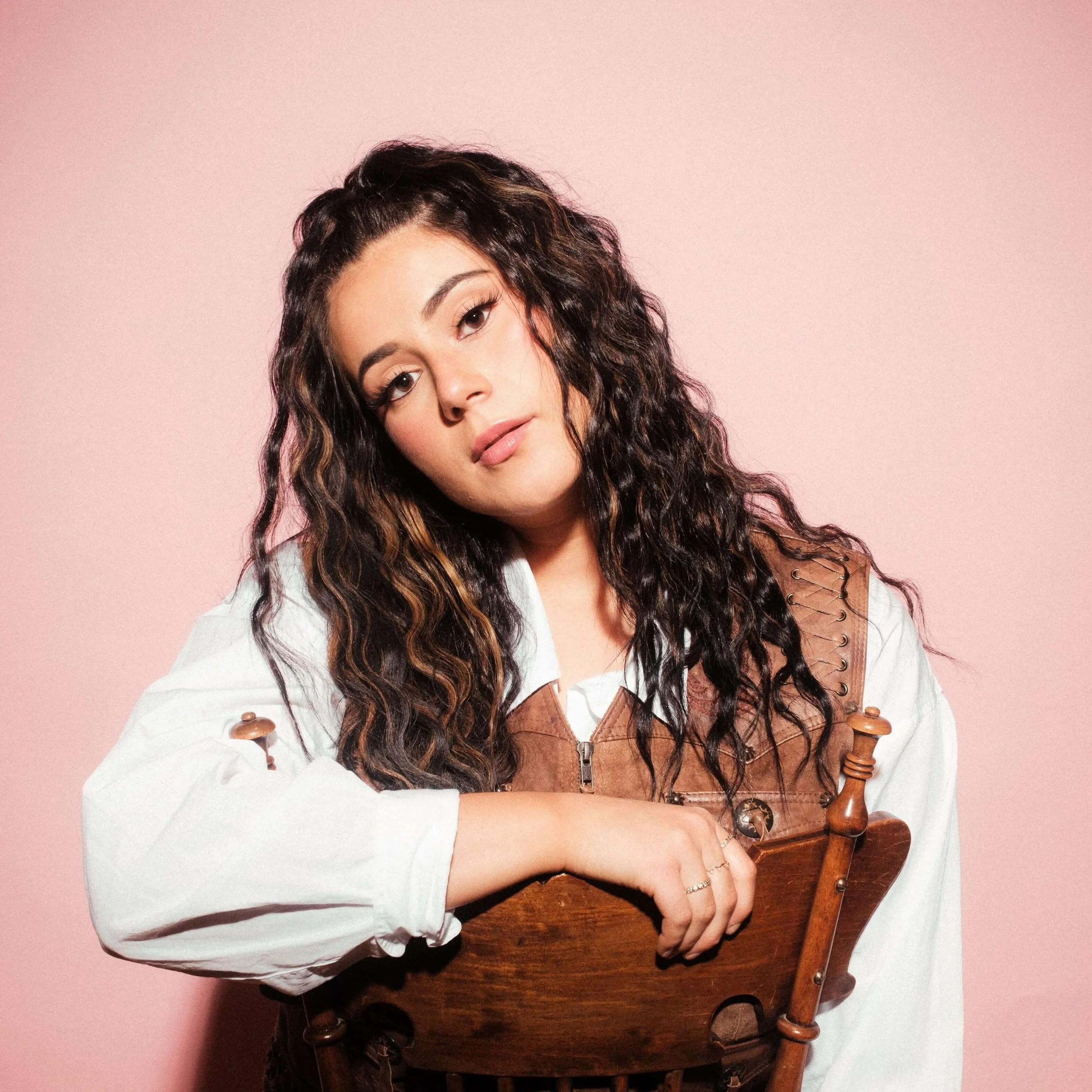 A woman with long curly dark hair, wearing a white shirt and brown leather vest, rests her arm on a wooden chair against a pink background.