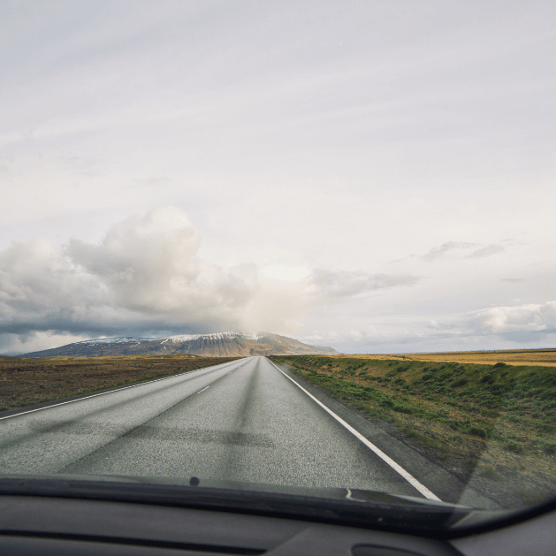 A view out of the windshield of a vehicle there are mountains in the distance and green and brown fields on either side of the road