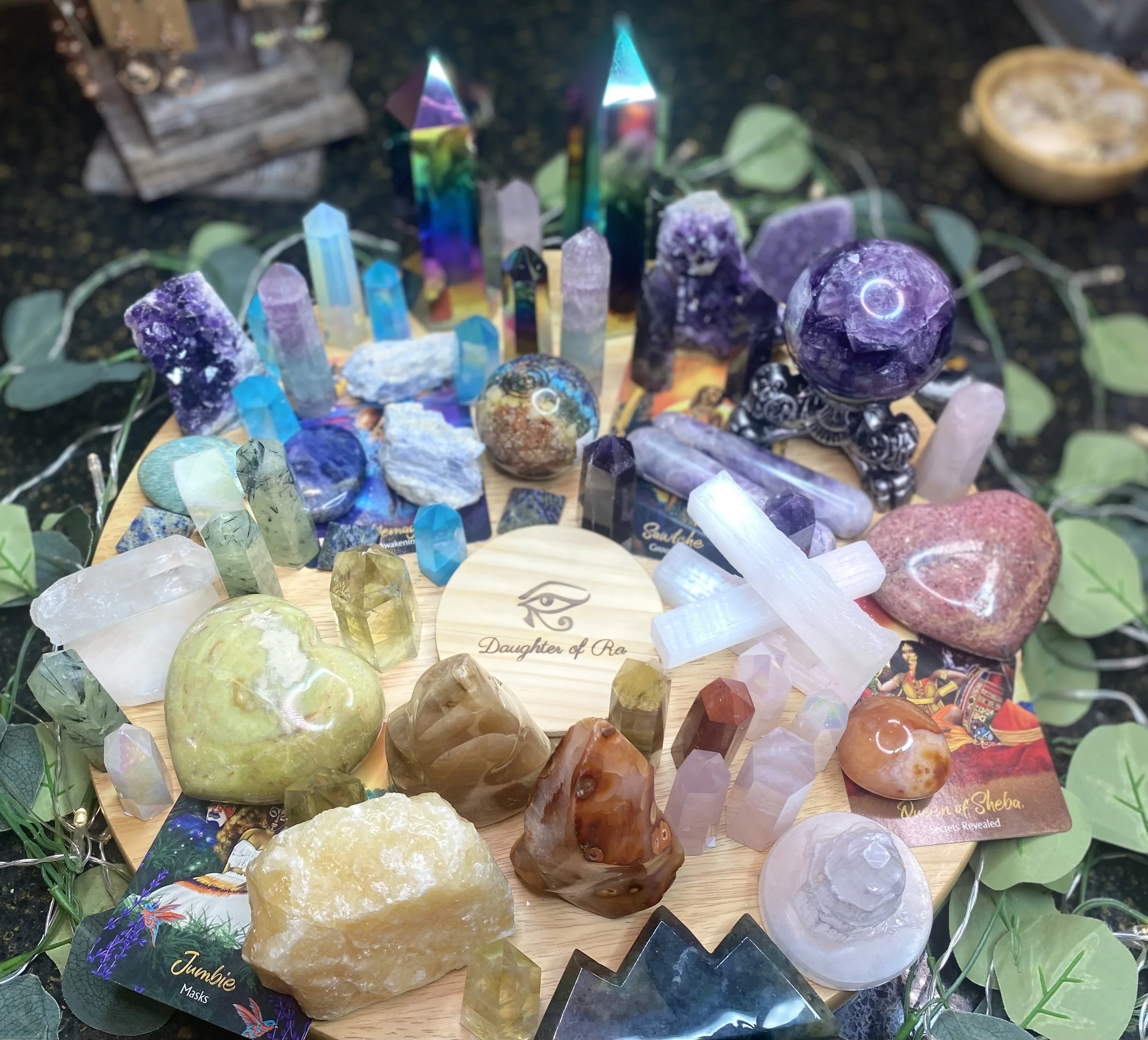 A circular wooden display filled with various colorful crystals and gemstones, including amethyst, quartz, selenite, and citrine. Some crystals are shaped into points, hearts, and spheres. The display is decorated with green leaves and includes a