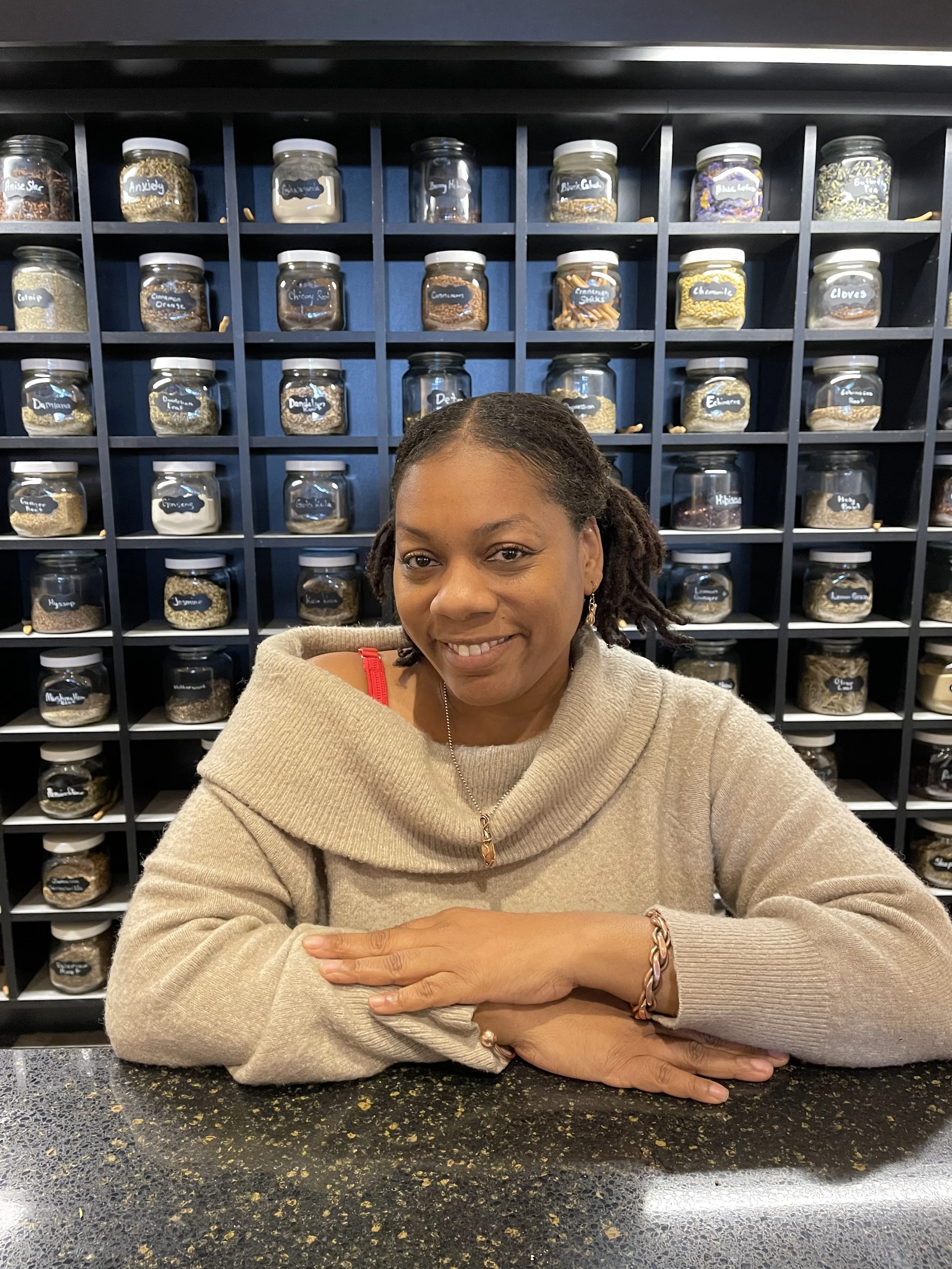 A woman with dreadlocks smiling, sitting at a counter with jarred spices and herbs behind her on a wall display.