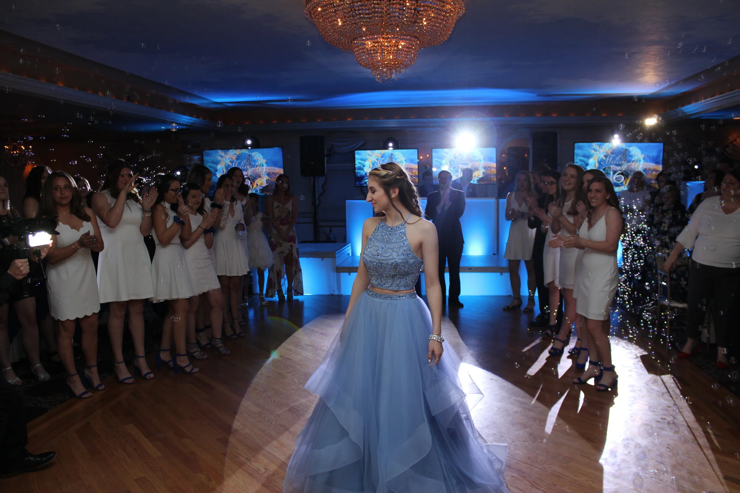 A young woman in a embellished blue dress is standing on a dance floor at a celebration. Surrounding her are people mostly dressed in white, watching and smiling. The room is decorated with a chandelier and multiple screens in the background displaying underwater scenes.