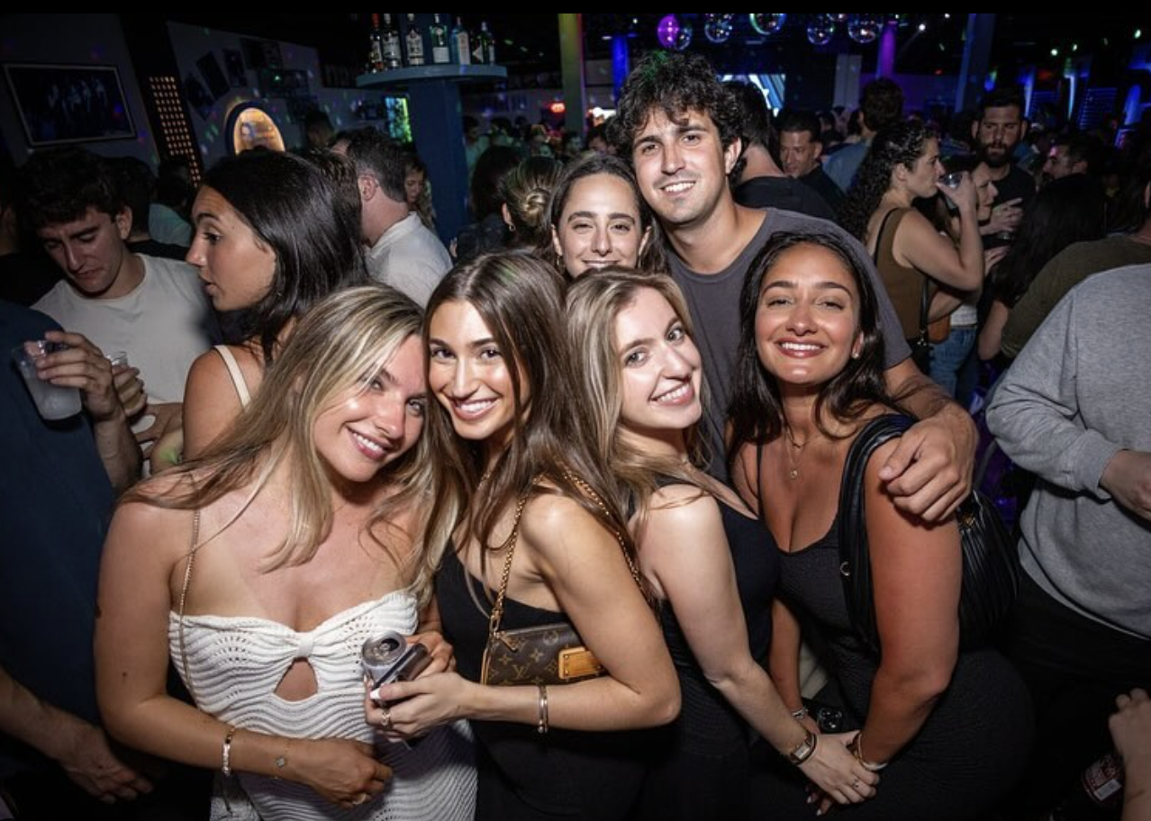 A group of six young adults, five women and one man, smiling and posing together at a lively nightclub with colorful lights and other people in the background.
