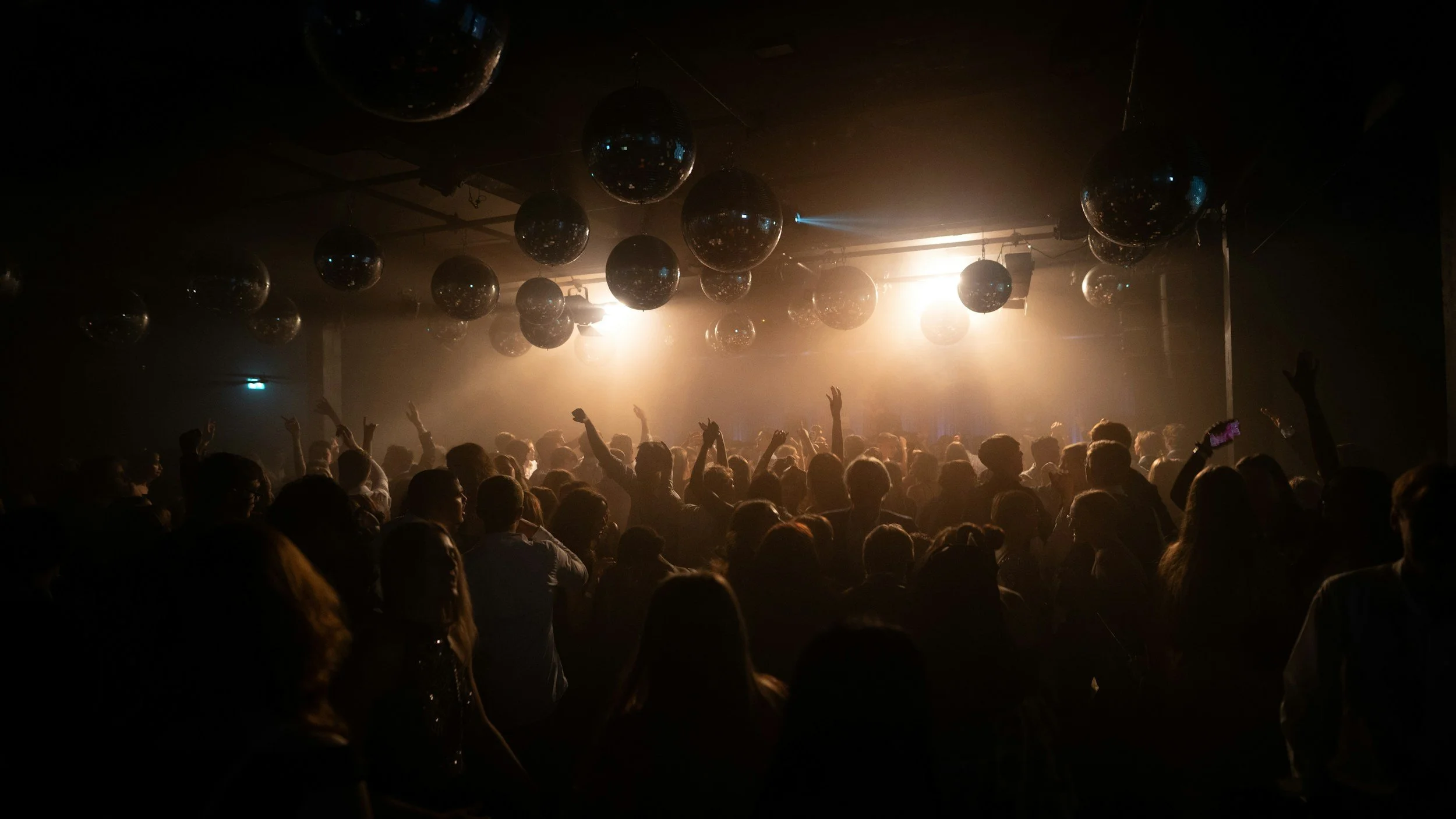 A large crowd of people dancing and enjoying a concert or nightclub with disco balls hanging from the ceiling and bright lights creating a lively atmosphere.