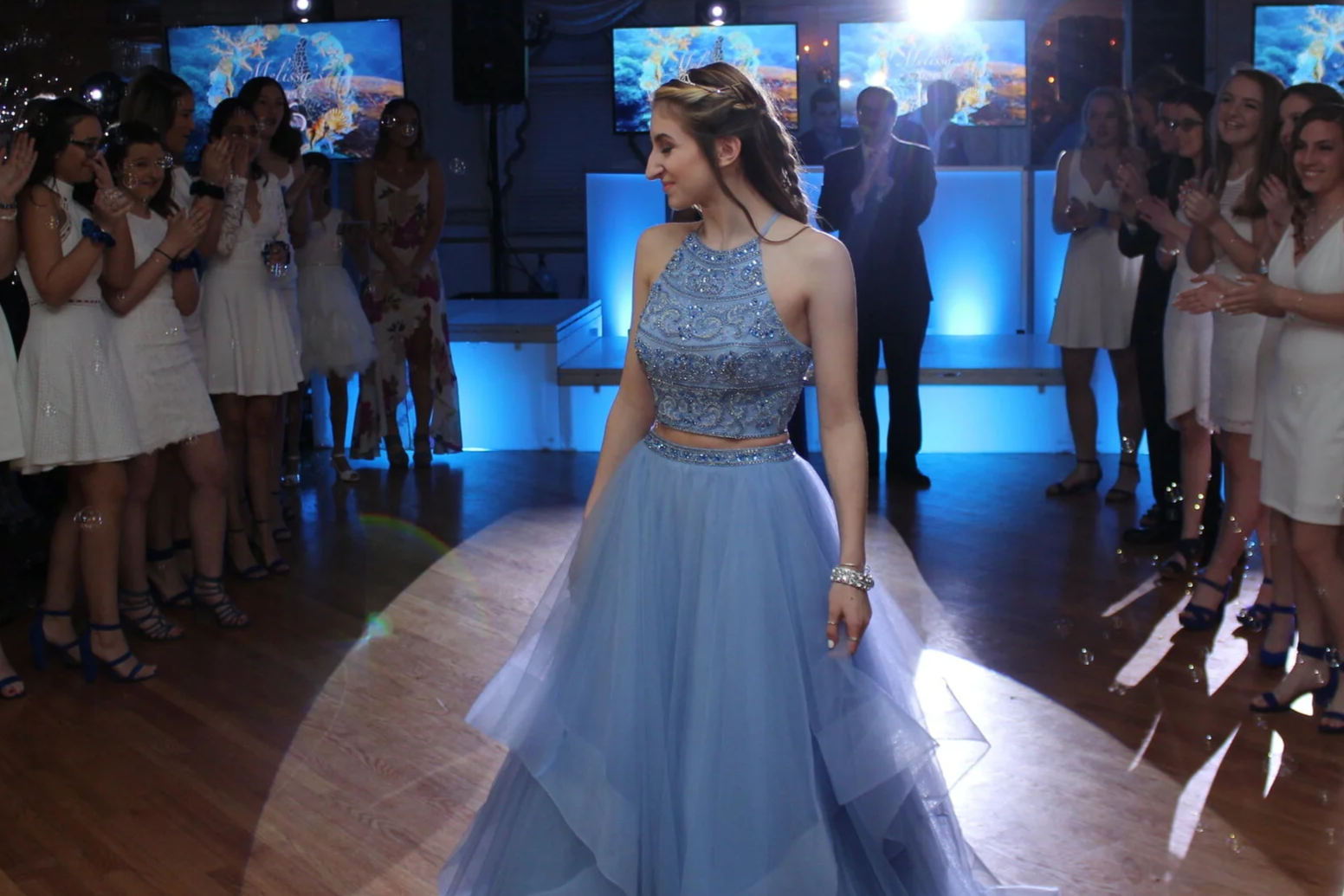 A young woman in a light blue, embellished dress is in the center of a dance floor, surrounded by onlookers clapping and smiling at her, in a dimly lit venue with blue lighting and screens displaying a seascape background.