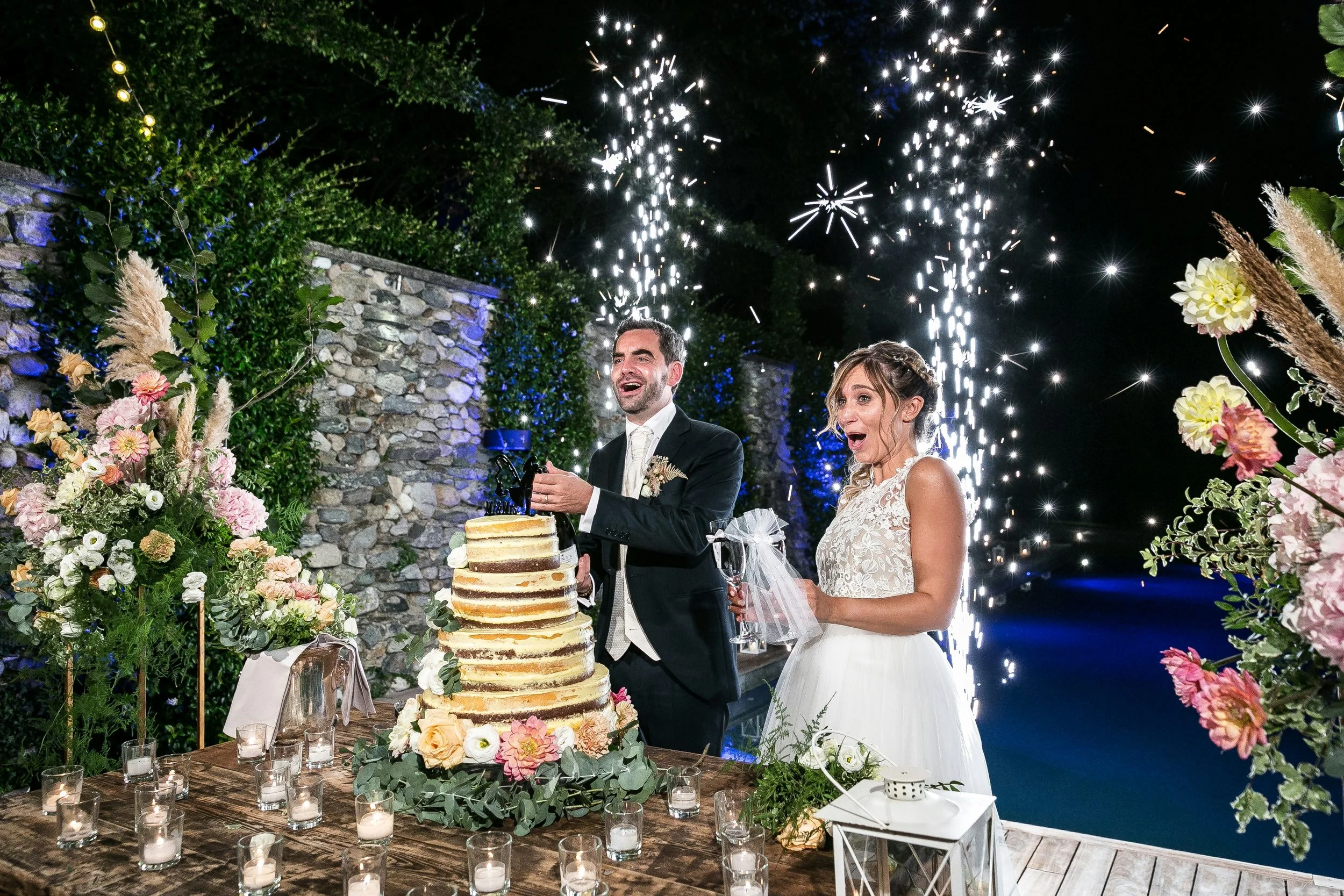 A bride and groom celebrate at a wedding reception, cutting a wedding cake outdoors at night. Fireworks light up the dark sky, and the scene is decorated with flowers and candles.