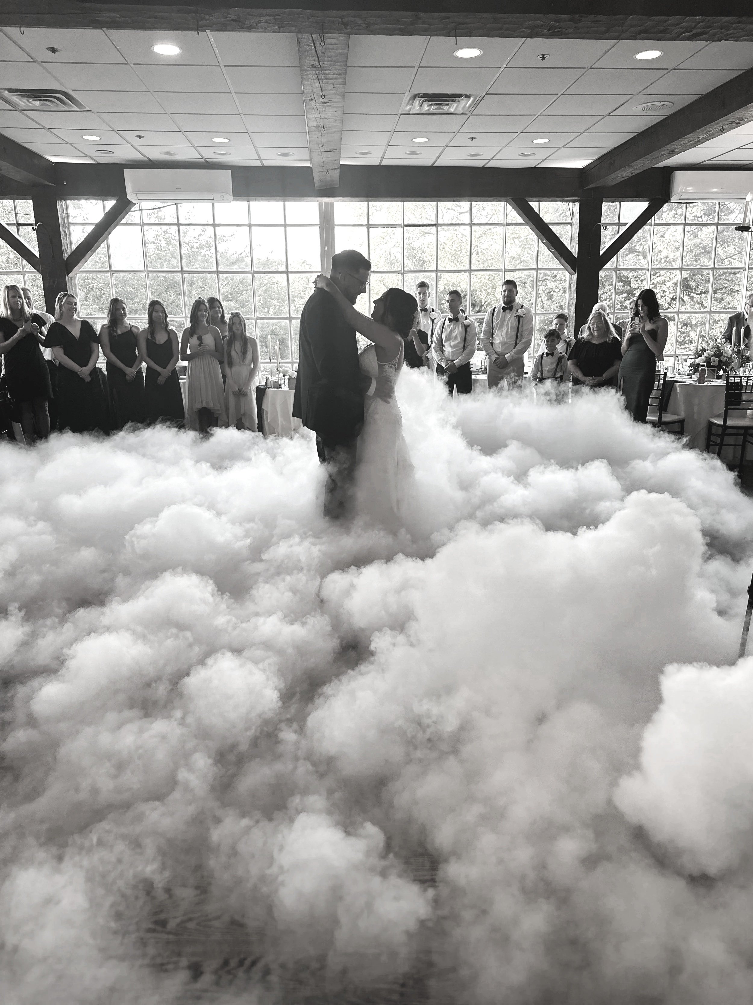 Black-and-white photo of a wedding reception where a bride and groom are dancing in a fog-filled dance floor, surrounded by guests watching and taking photos inside a venue with large windows.