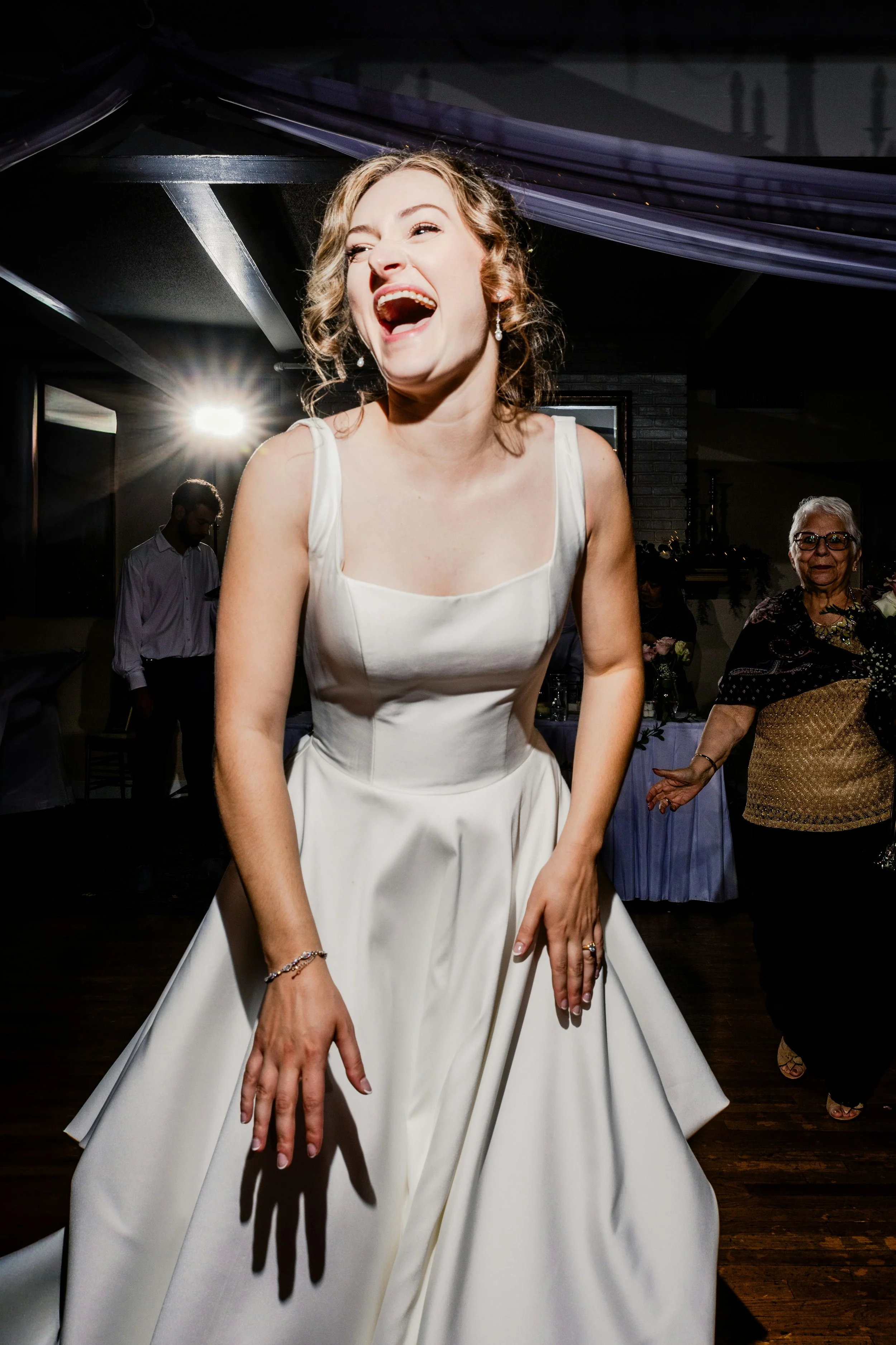 A woman in a white wedding dress at a celebration or wedding reception, smiling and laughing with her hands in front of her, in a dimly lit indoor setting with decorations and other guests in the background.