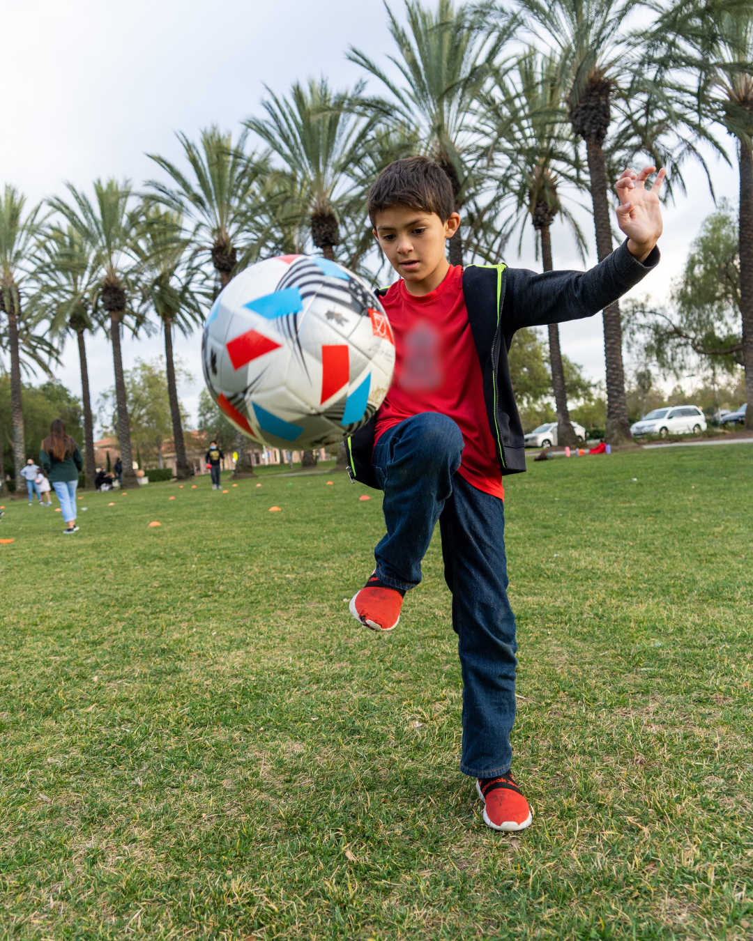 A young boy playing soccer outdoors on a grassy field, with palm trees and other people in the background.