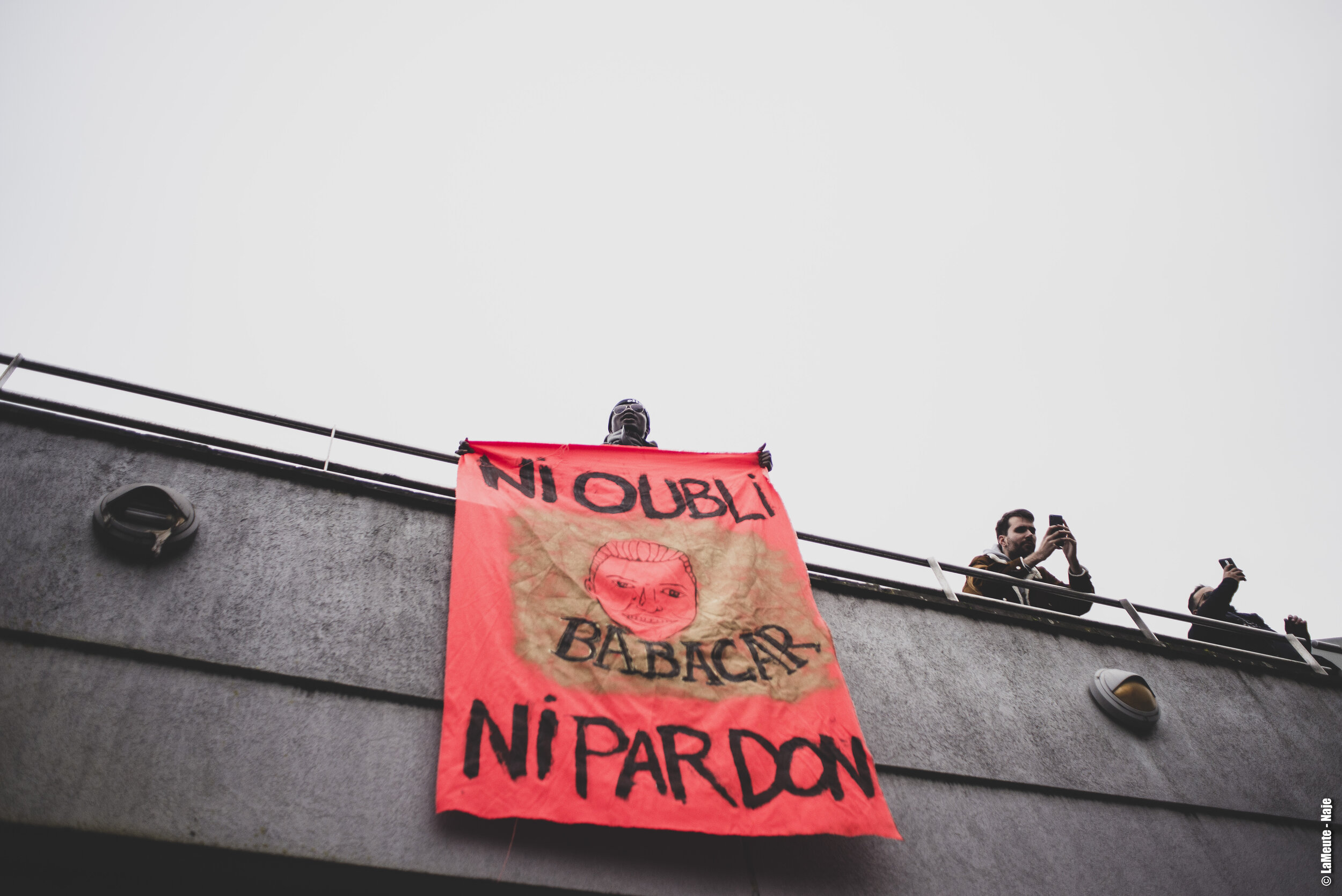   Sur une passerelle au dessus du parking, et alors qu’une nouvelle prise de parole s’organise, un manifestant déploie une banderole.  ©LaMeute - Naje 