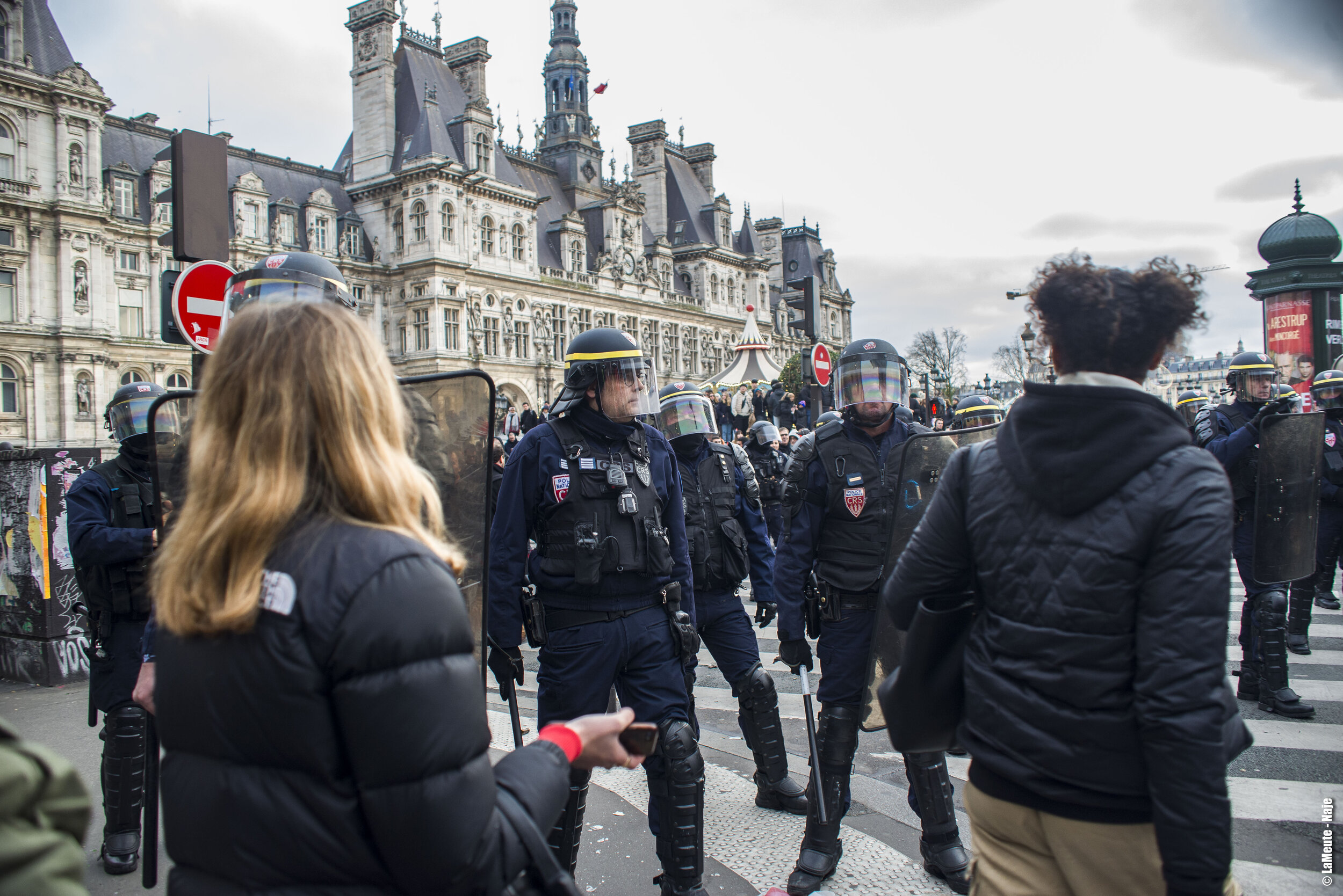   Devant l’hôtel de la Mairie de Paris, le dispositif policier est très fourni. De part et d’autres manifestant•es et touristes s’en amusent et échangent parfois quelques mots tandis que le cortège poursuit son chemin.  ©LaMeute - Naje.      