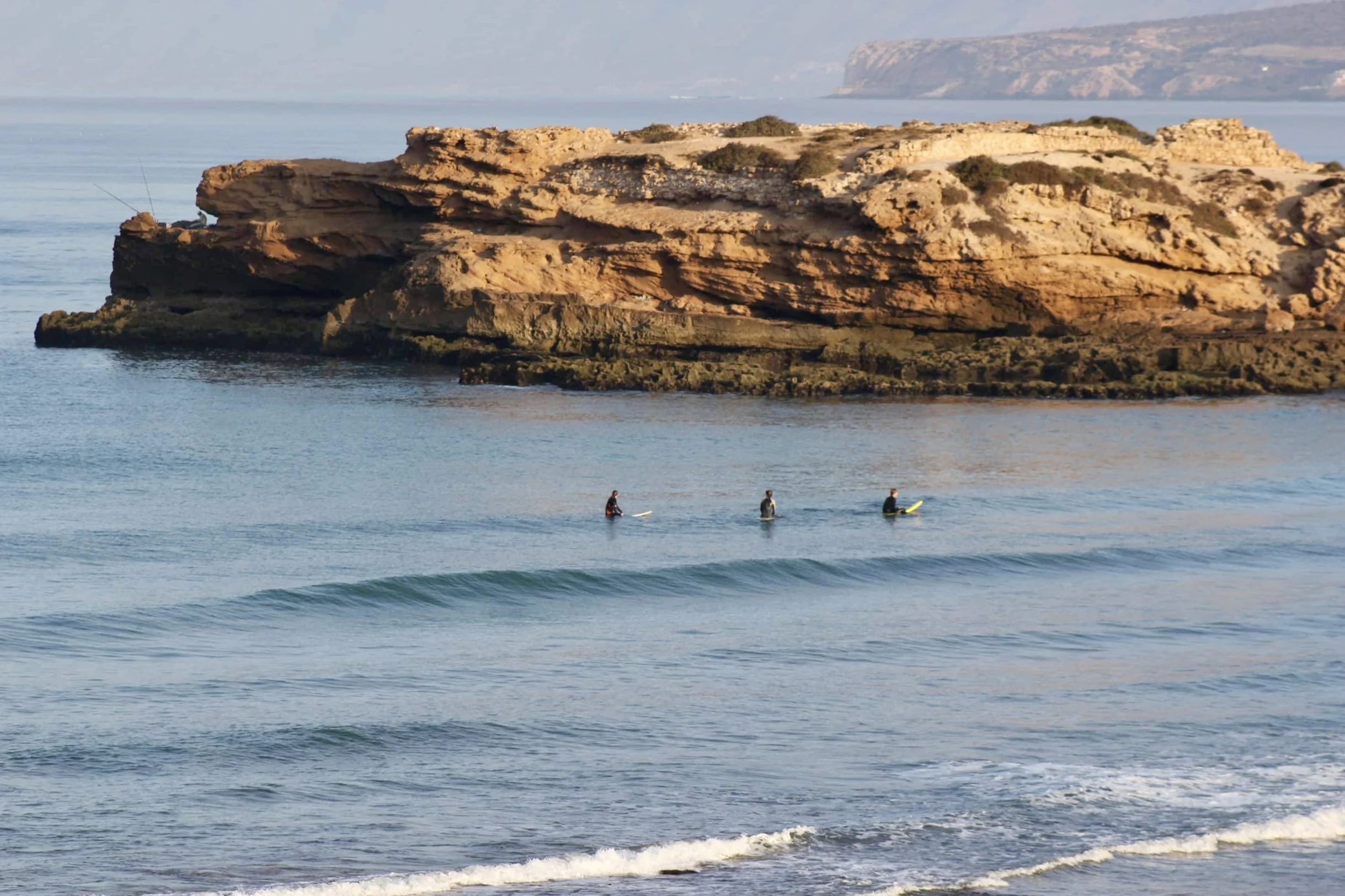 Three surfers waiting in the lineup at Devil's Rock surf spot in Tamraght, Taghazout Bay, Morocco, with gentle waves and coastal rock formations in the background.