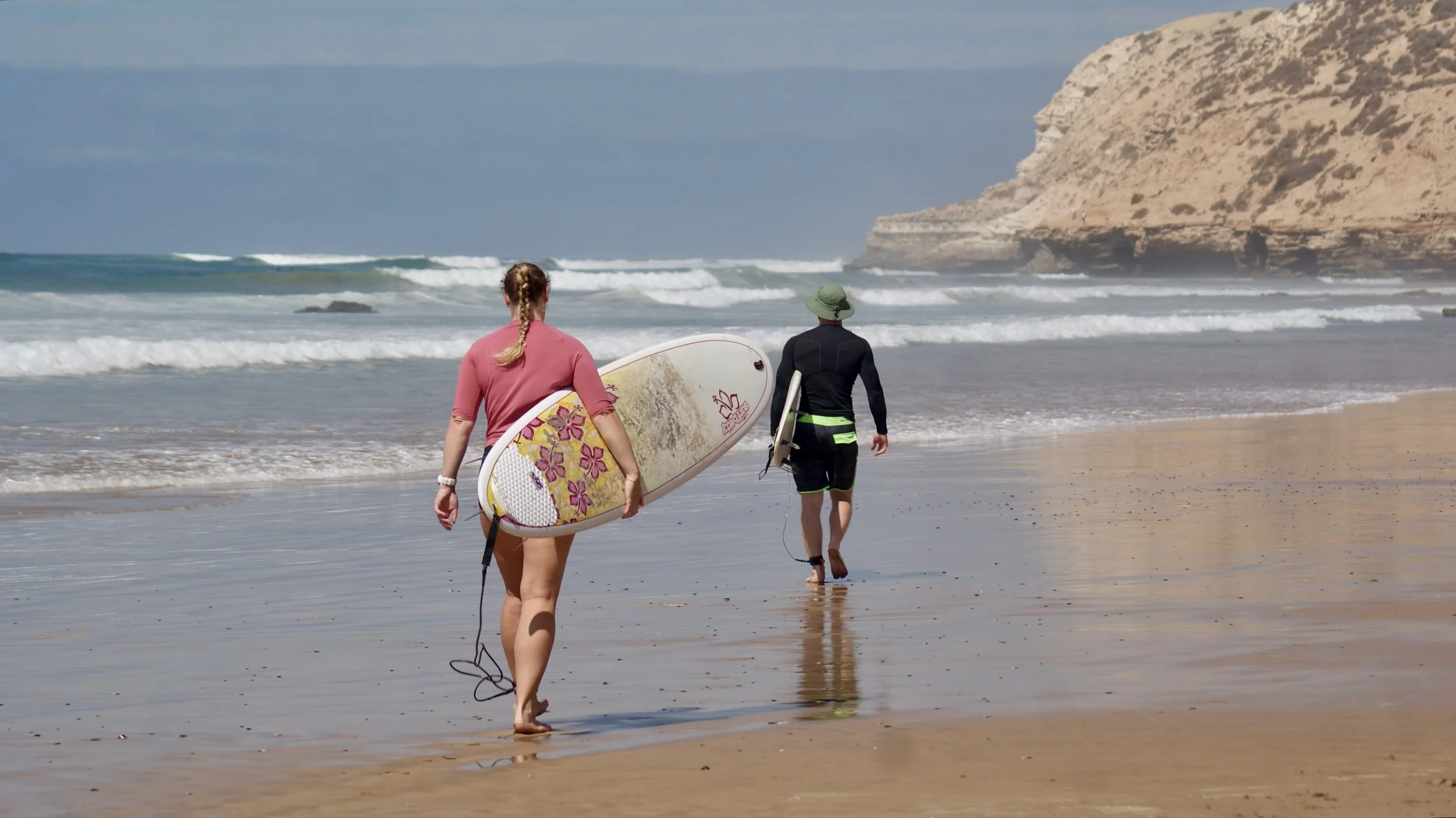 Two surfers walking along the beach towards Mysteries surf spot of Taghazout, Morocco, with waves breaking along the coastline.