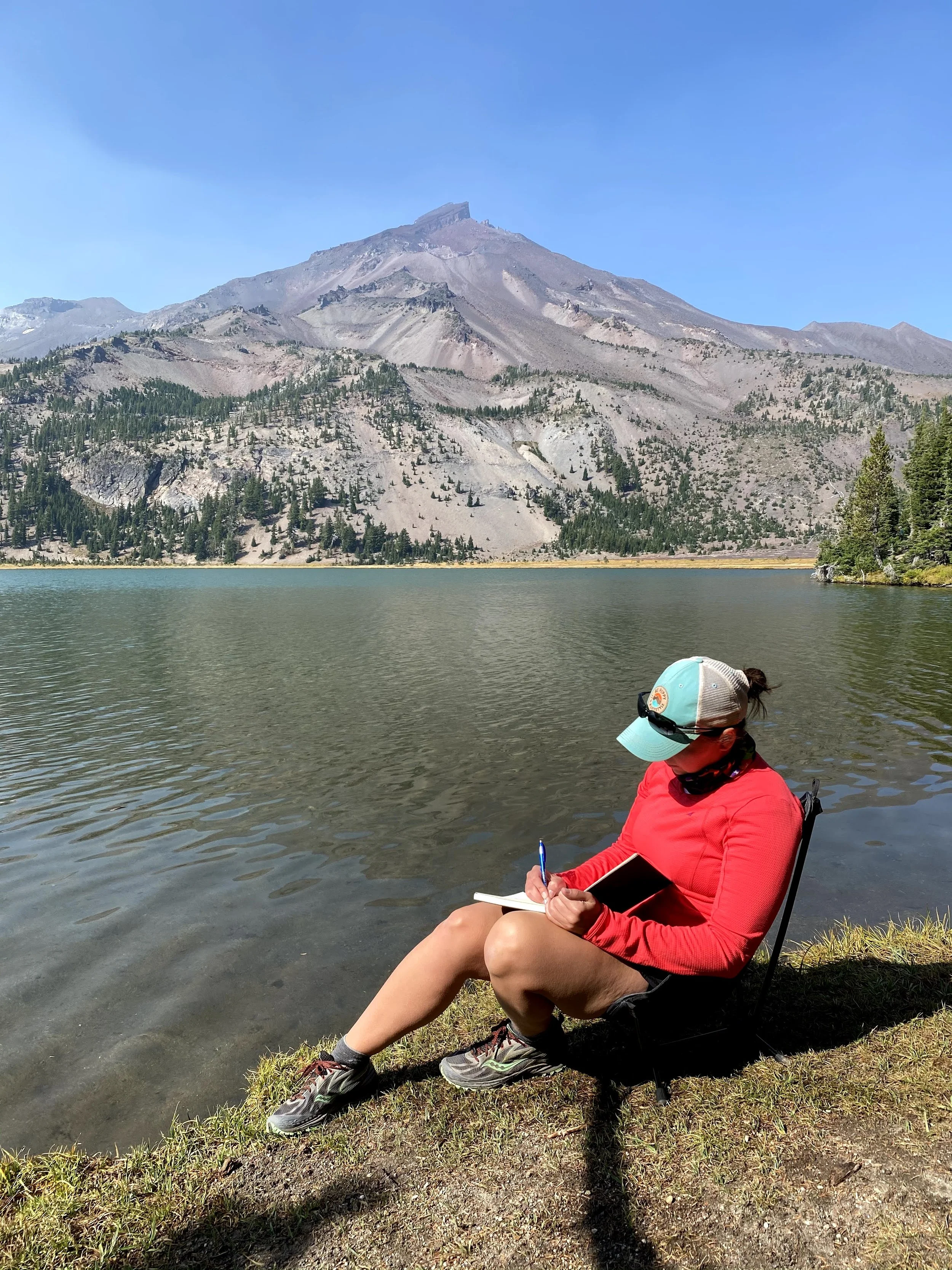 Coach Jenn sitting next to lake in a low chair, with mountain in background. she is taking notes in a journal.