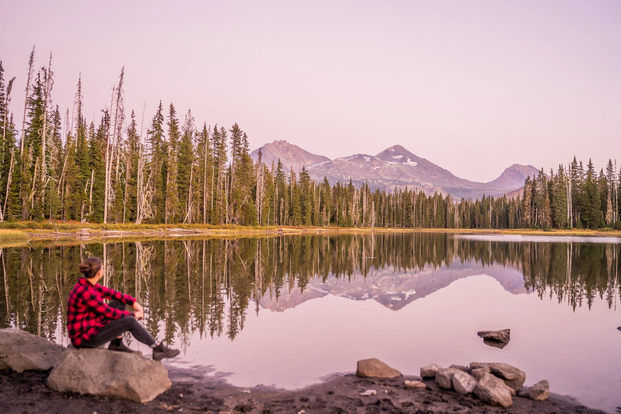 Coach Jenn, sitting lake side at sunset, with three sisters mountain in background and reflecting in lake. She is wearing red plaid shirt, jeans, and whole picture has a pink overtone