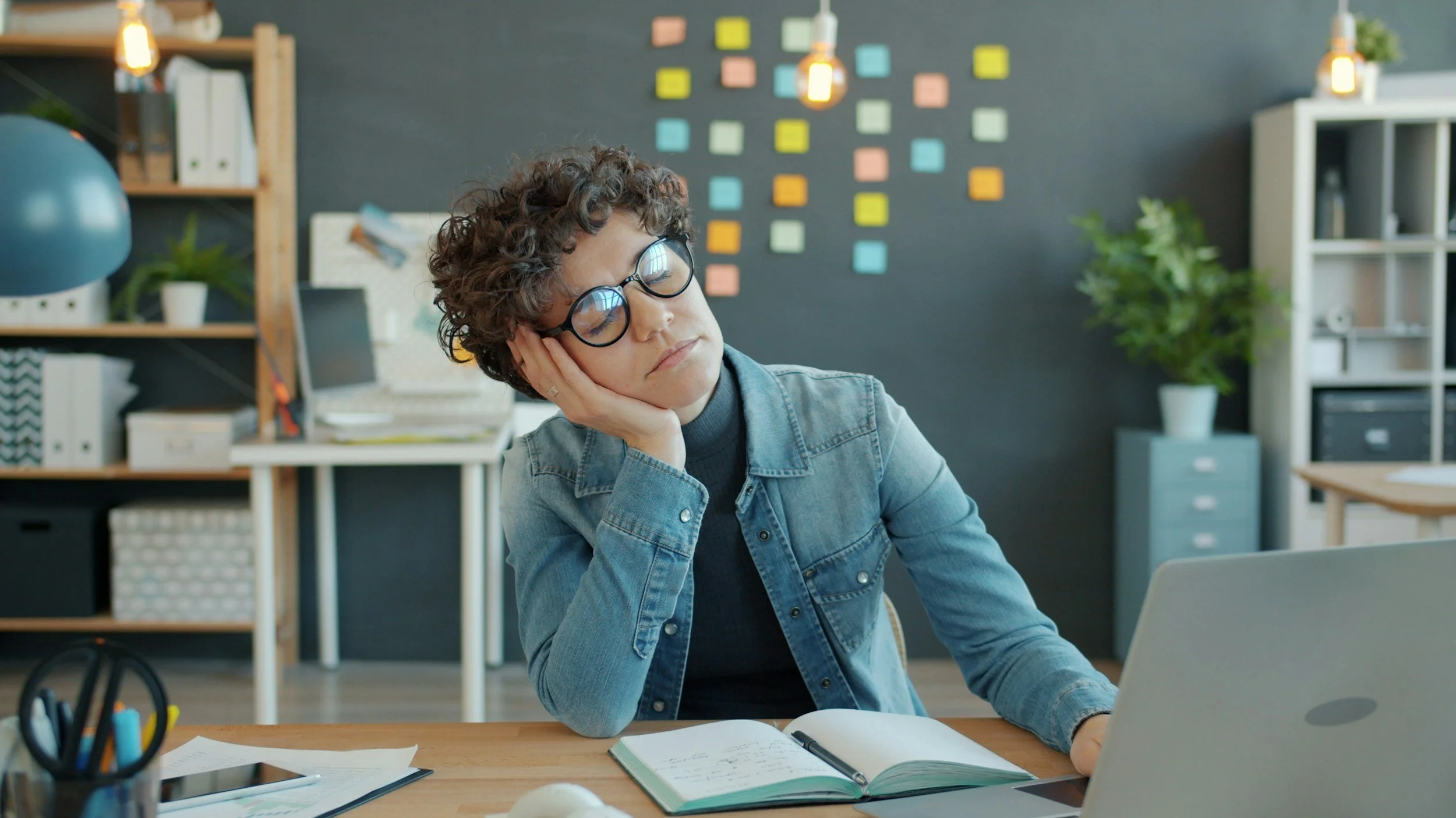 woman in jean jacket at computer looking burnt out, post it notes on wall behind