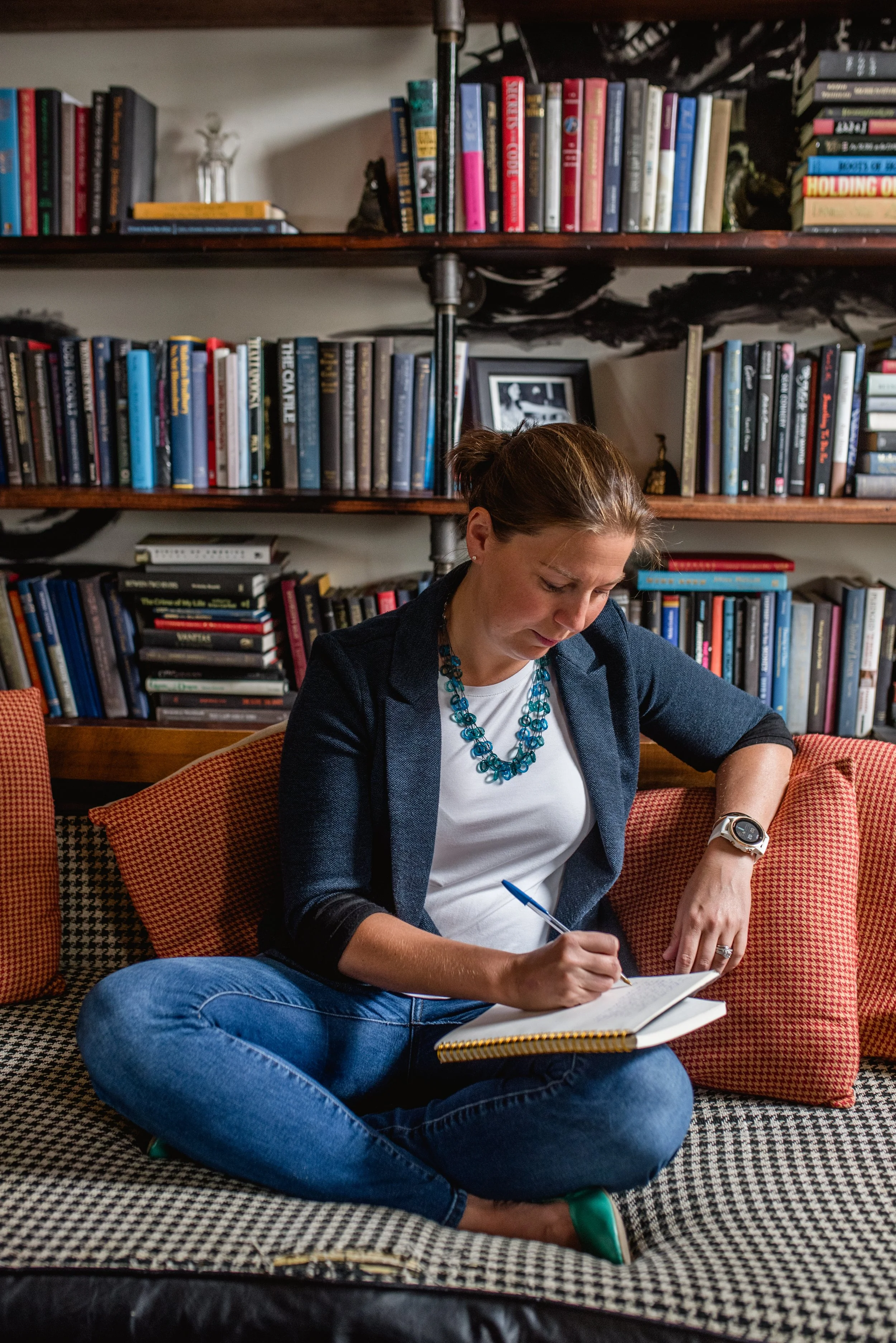 coach jenn masse journaling. she is on an orange couch with book shelves behind her. she is wearing a teal blazer, white shirt, and blue jeans.
