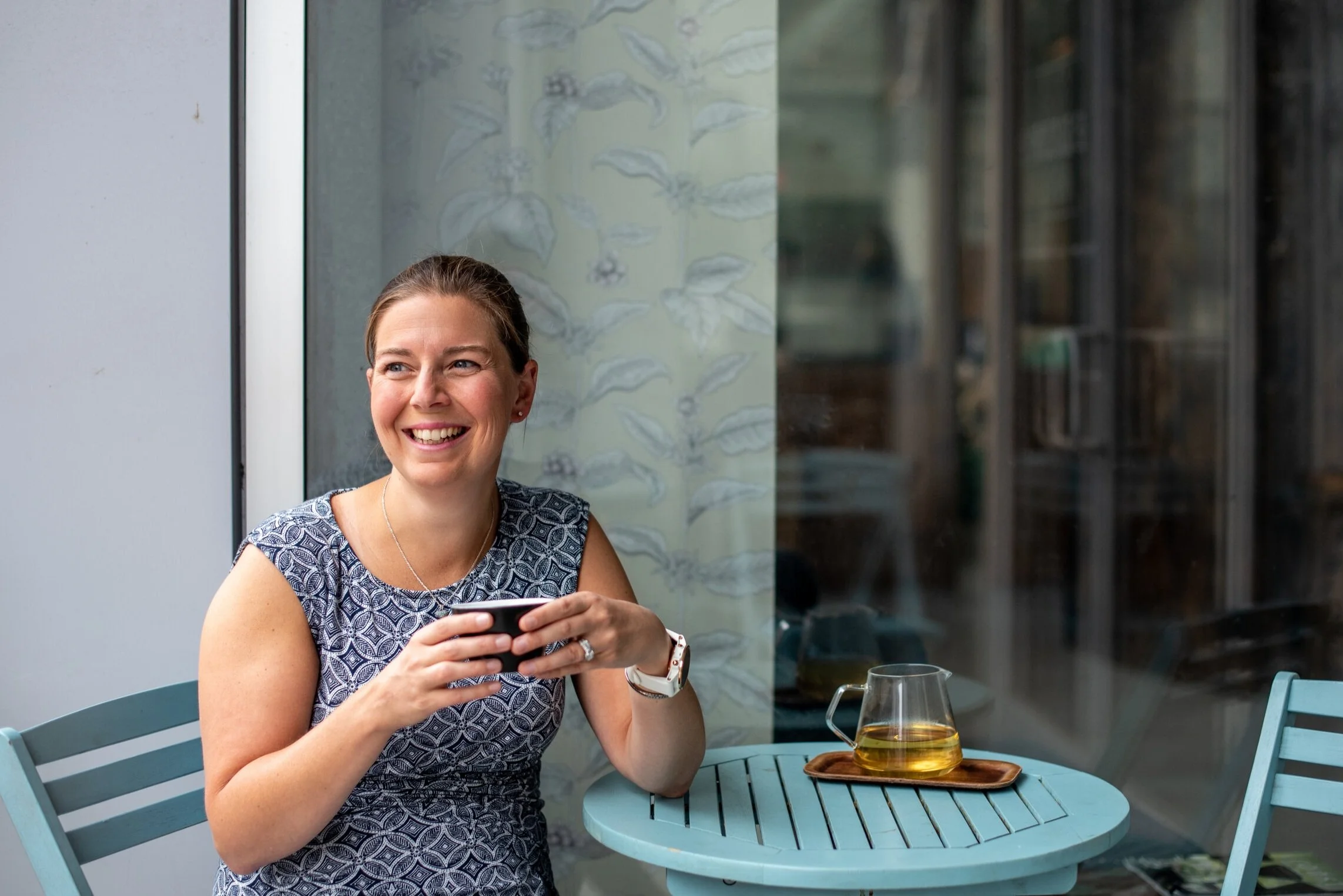 Jenn Masse, holding cup of tea, in blue dress at outdoor cafe