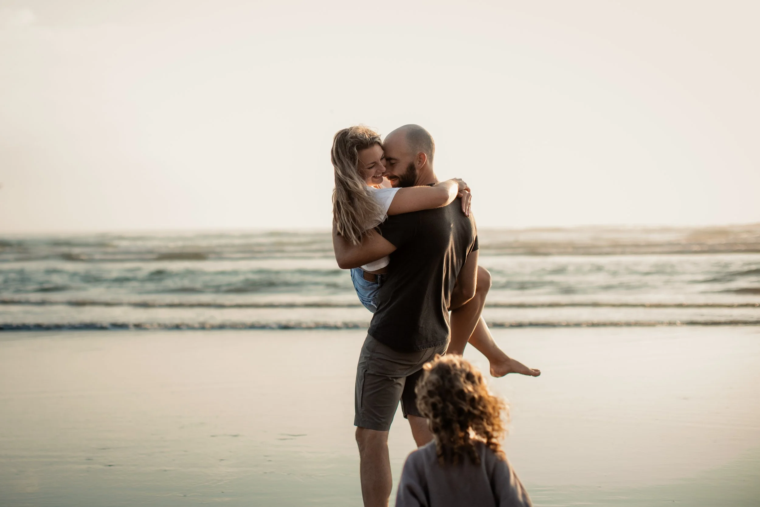 father holding mother while being chased by toddler on the beach at sunset during a family photoshoot