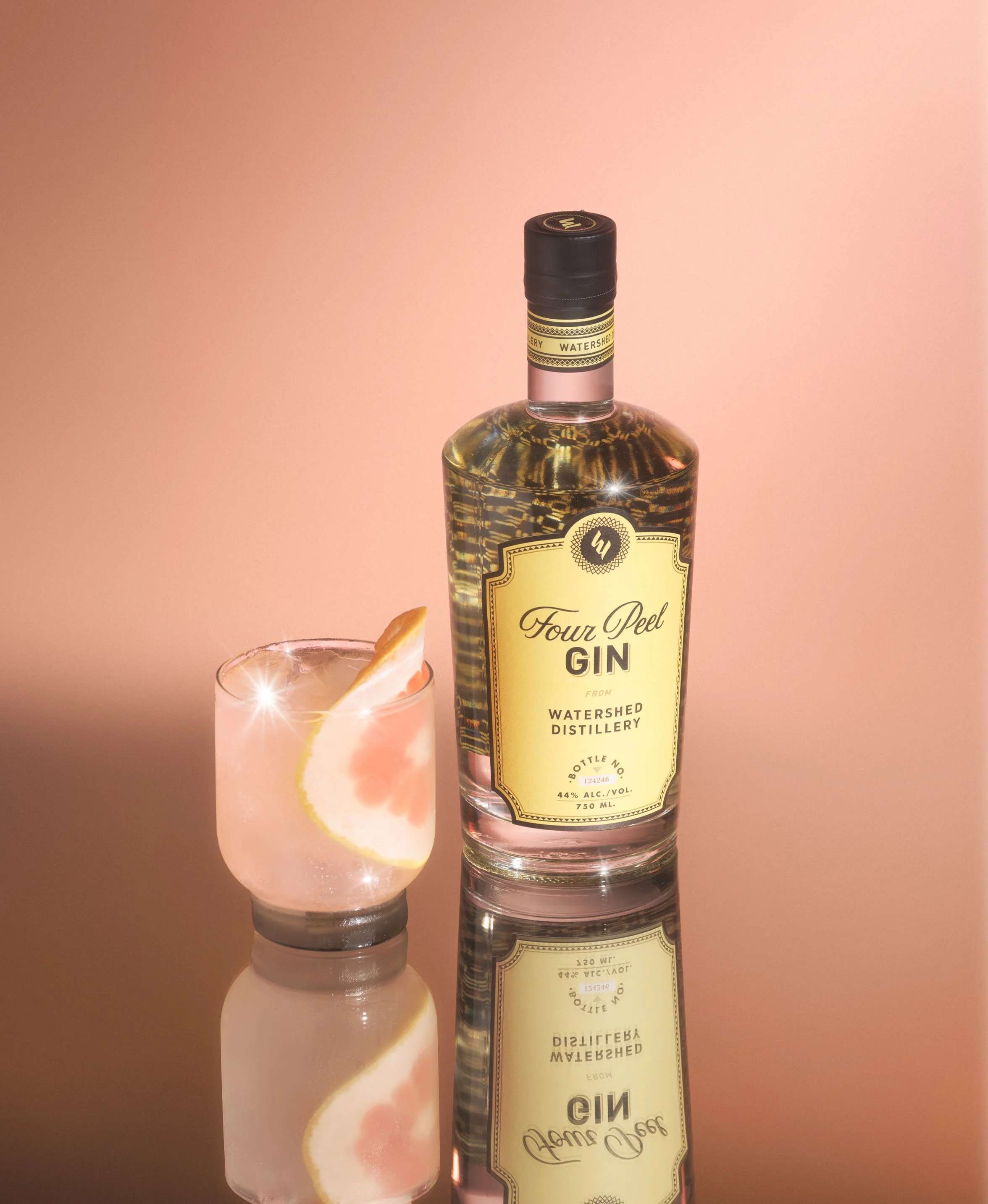 A bottle of Four Peel Gin from Watershed Distillery next to a glass filled with a light-colored cocktail garnished with a lemon slice on a reflective surface with a pink background.