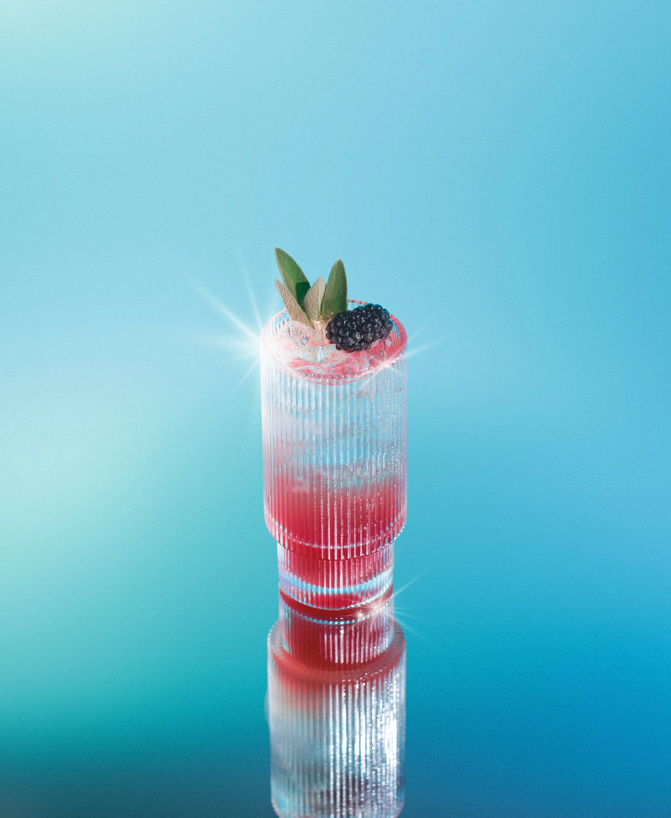 A tall glass with a pink drink, garnished with green leaves and a blackberry, on a reflective surface against a light blue background.