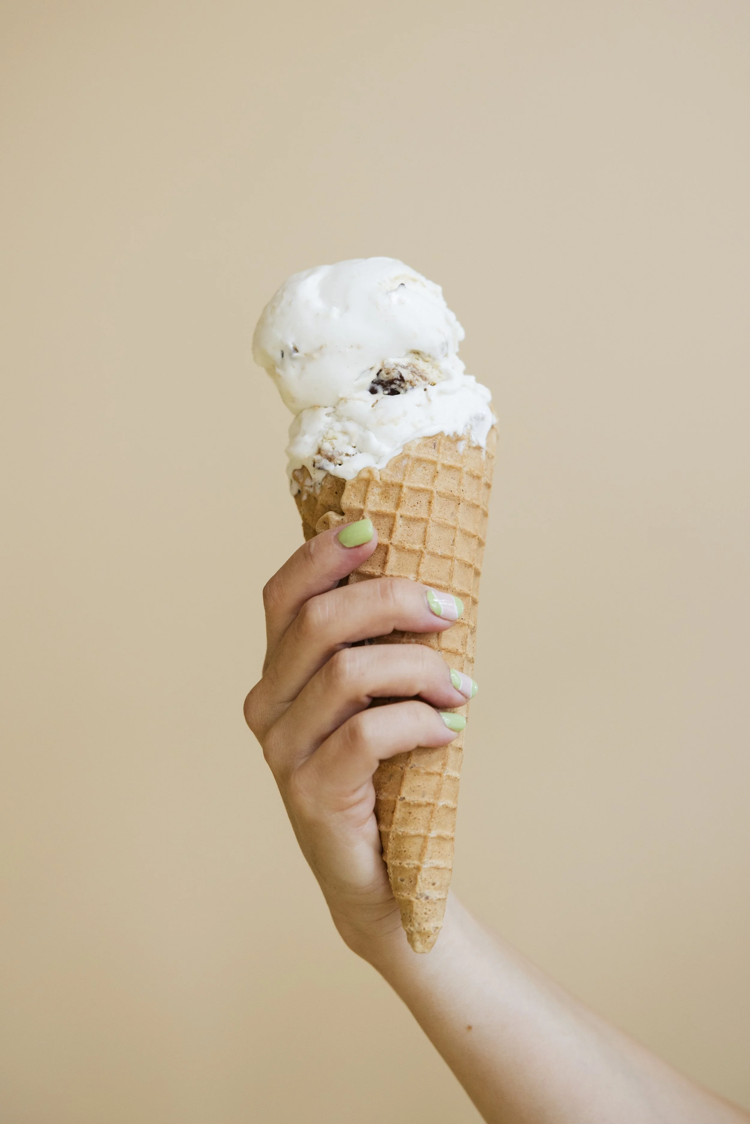 Hand holding a waffle cone topped with scoops of white ice cream and chocolate pieces