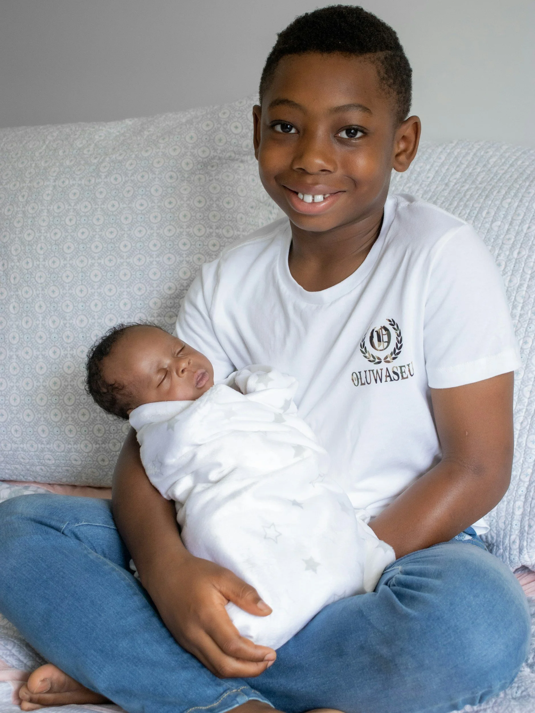 A young boy with a big smile, wearing a white shirt and jeans, sitting on a bed and holding a sleeping newborn baby wrapped in a white blanket.
