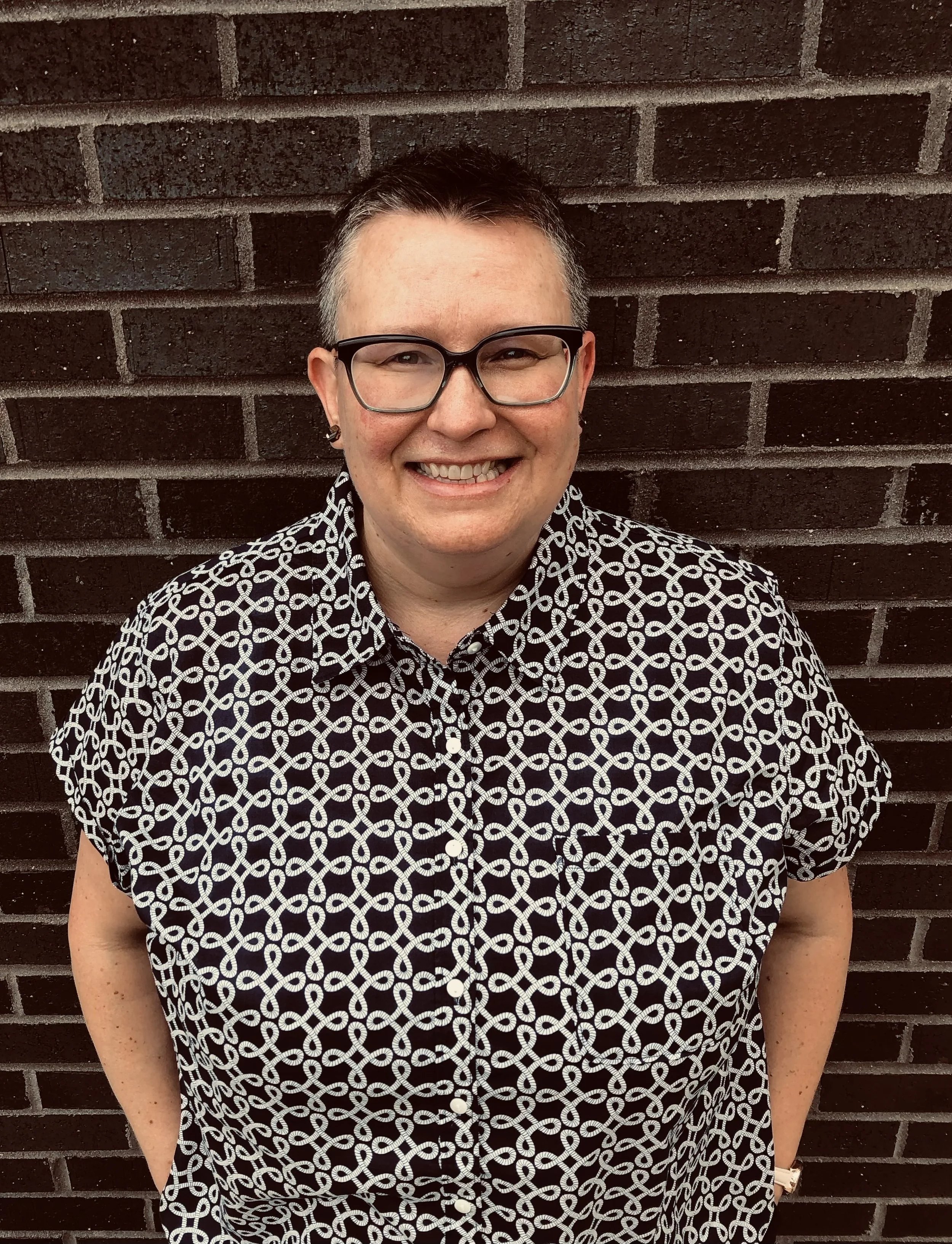 [A white woman in her mid-40s stands in front of a brick wall. She has short, brown/gray hair, hazel eyes, blue glasses, and small wooden stud earrings. She is smiling and wearing a blue and white patterned collared shirt.