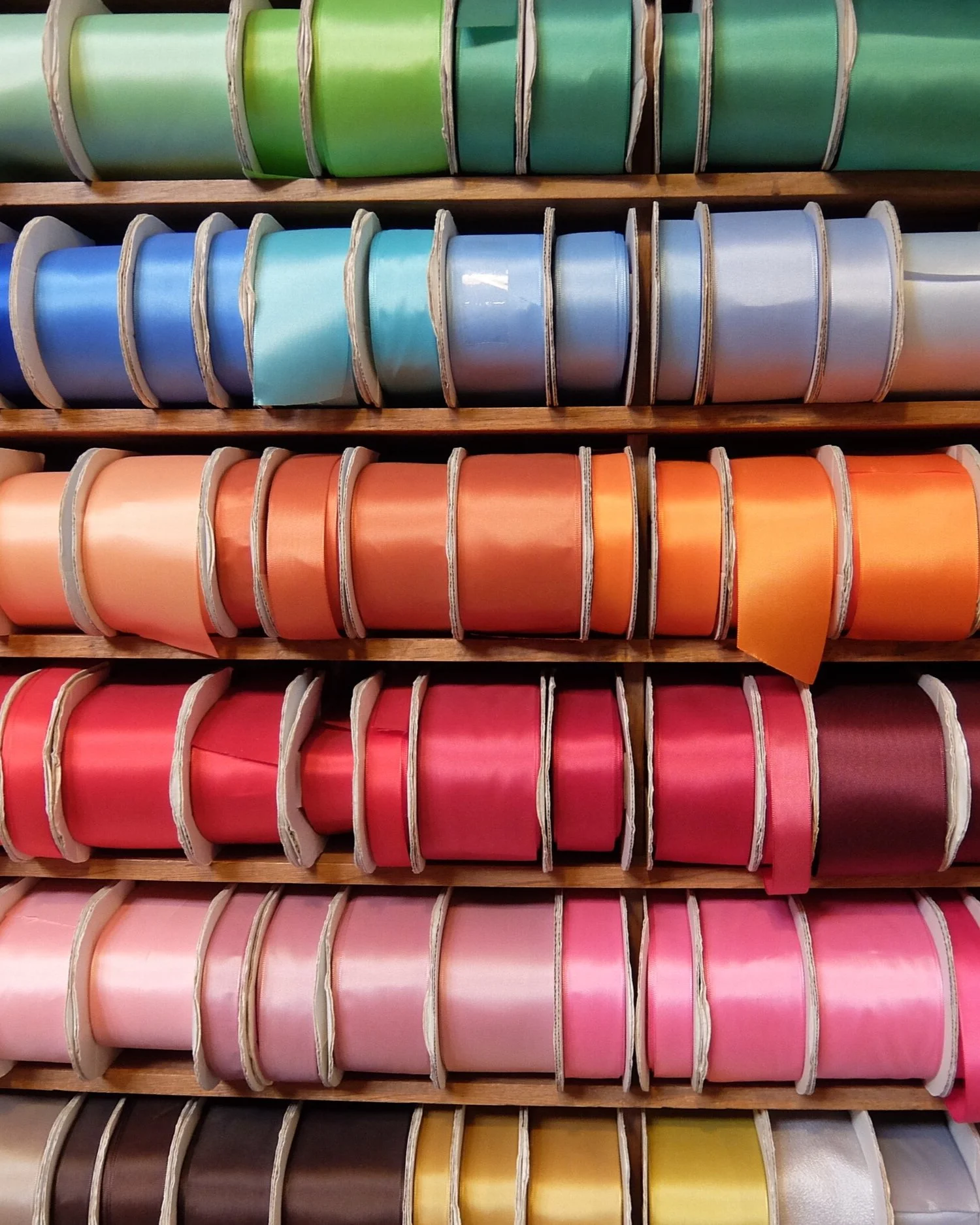 Multiple spools of satin ribbon in various colors, arranged on wooden shelves in a store.
