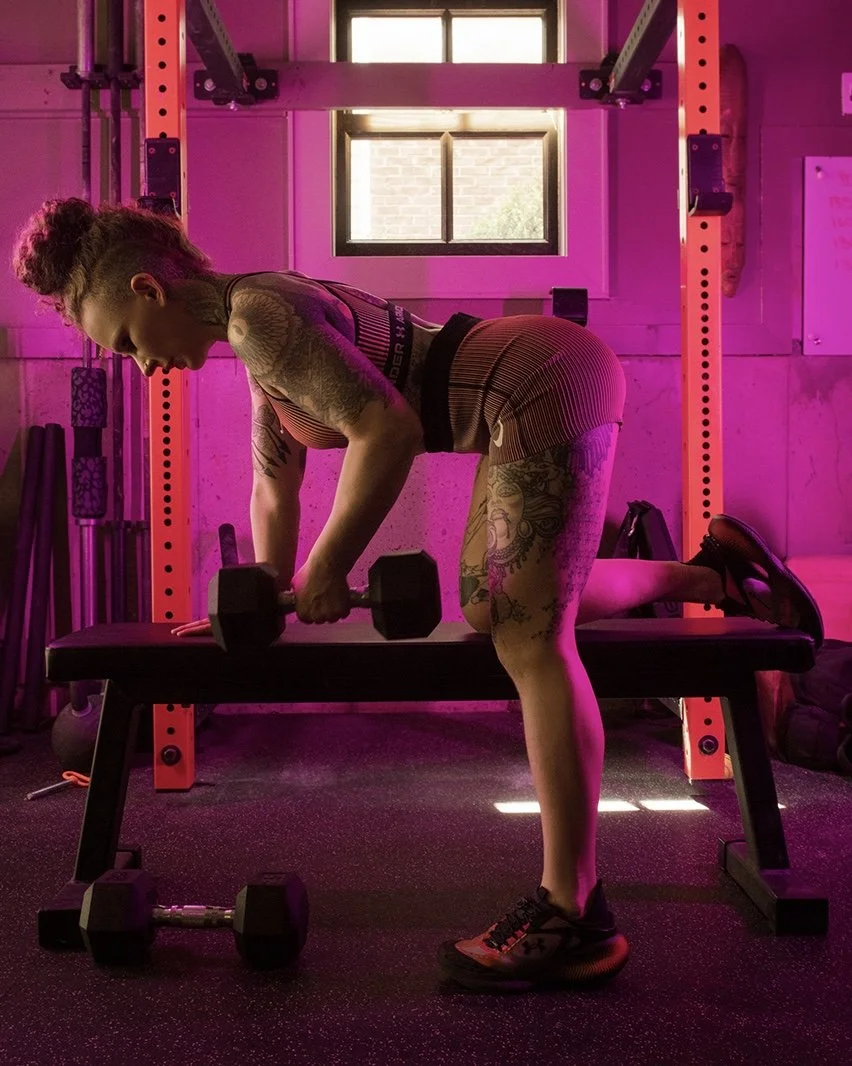A woman with tattoos in a gym, performing a workout with dumbbells on a bench, illuminated by pink lighting.