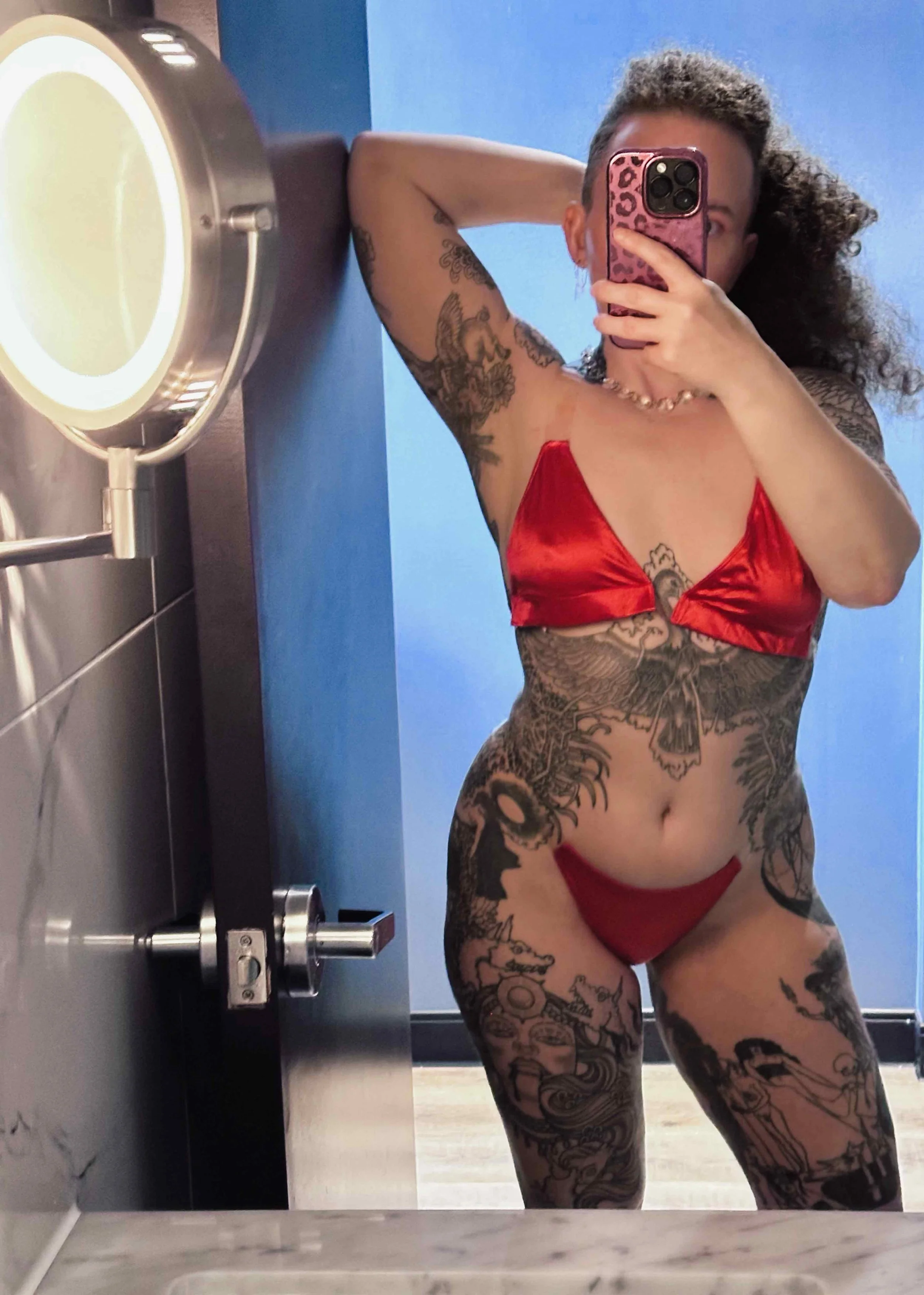 Person with tattoos wearing a red satin crop top and matching underwear taking a mirror selfie in a bathroom.