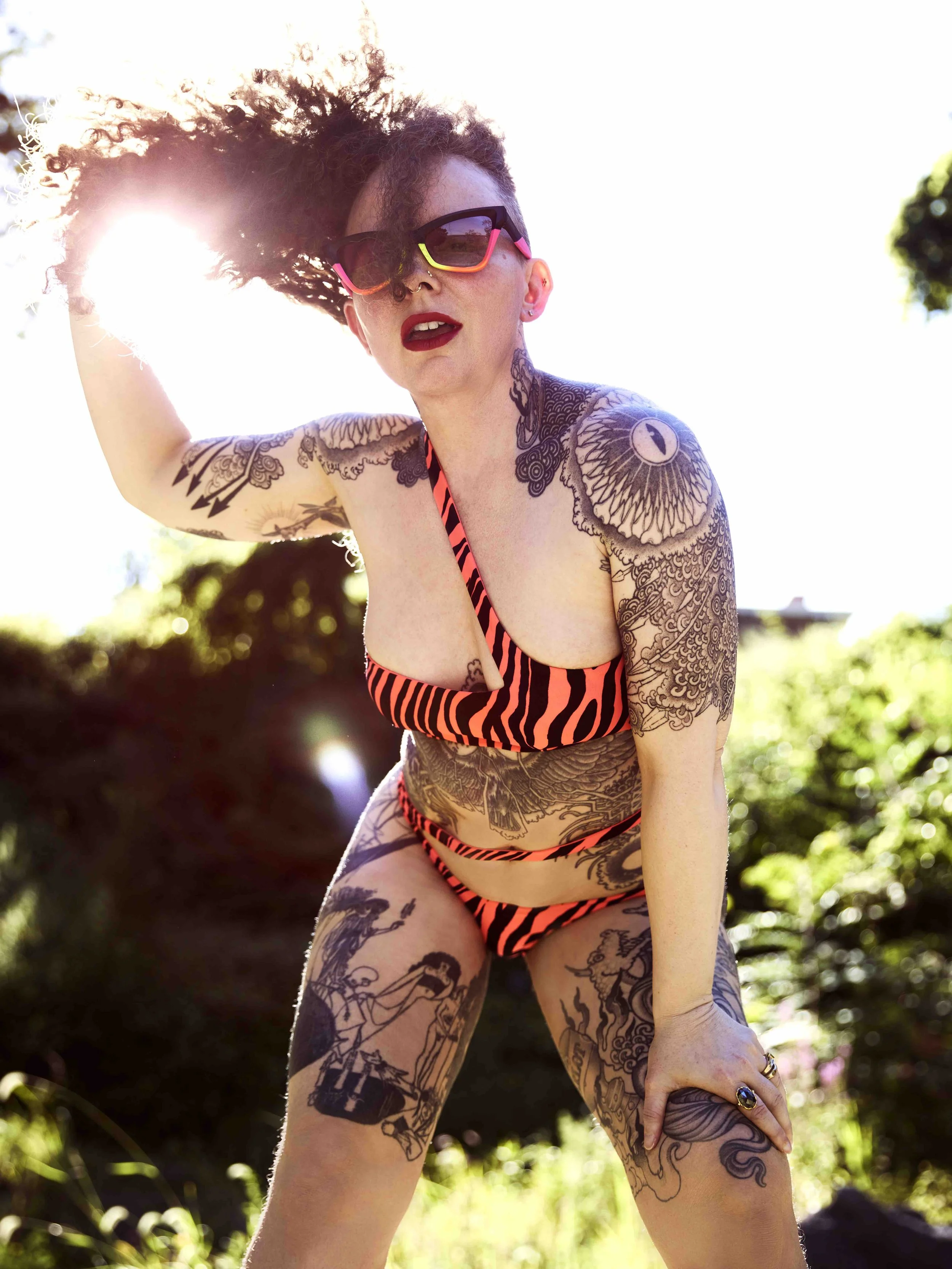 An alternative model with curly hair, wearing sunglasses and a tiger striped bikini, outdoors in sunlight, with visible tattoos on her arms, torso, and legs.