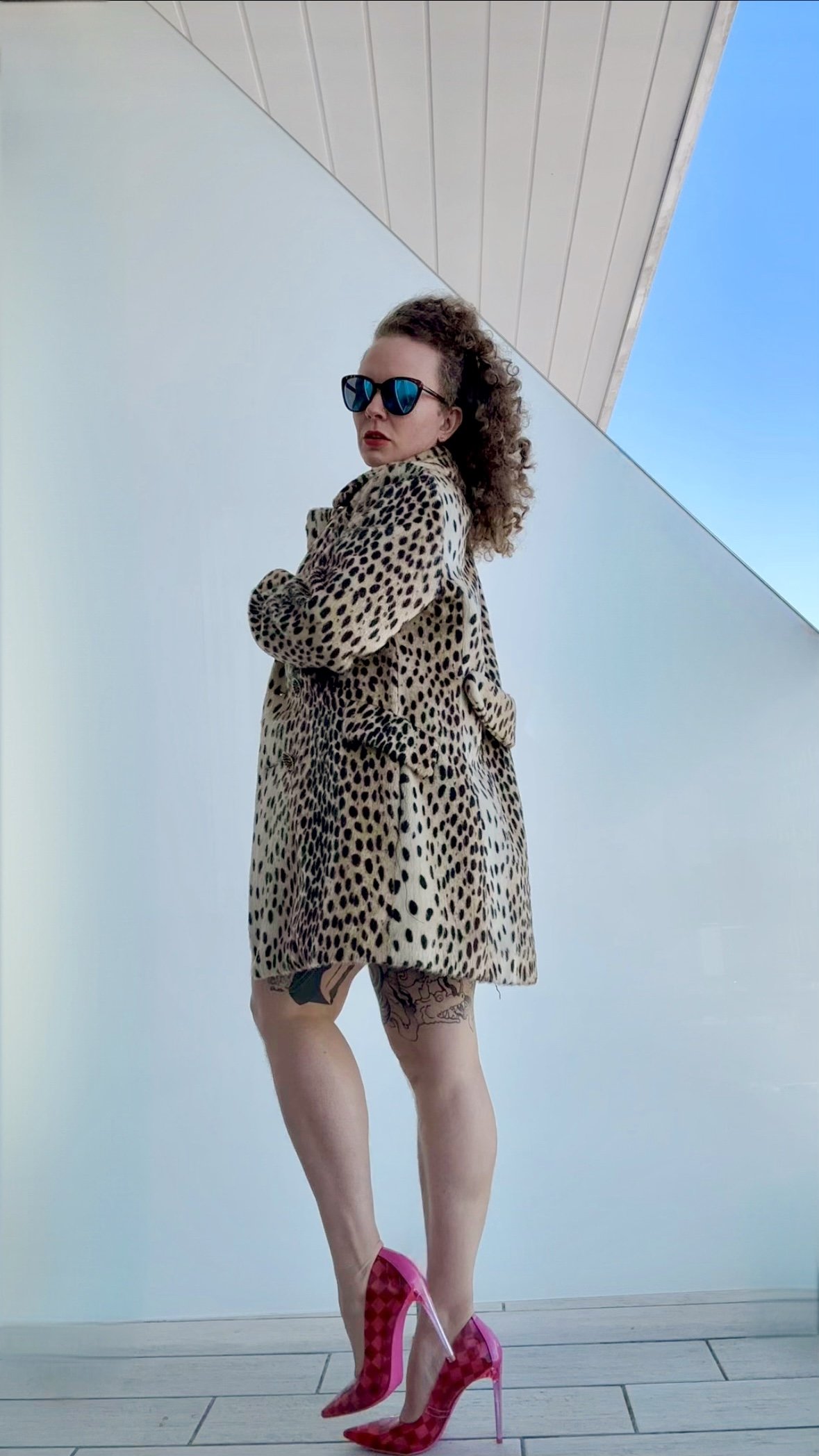 A woman with curly hair wearing dark sunglasses, a leopard print coat, pink high heels, and black tattoos on her legs, standing against a modern building with white walls and a blue sky.