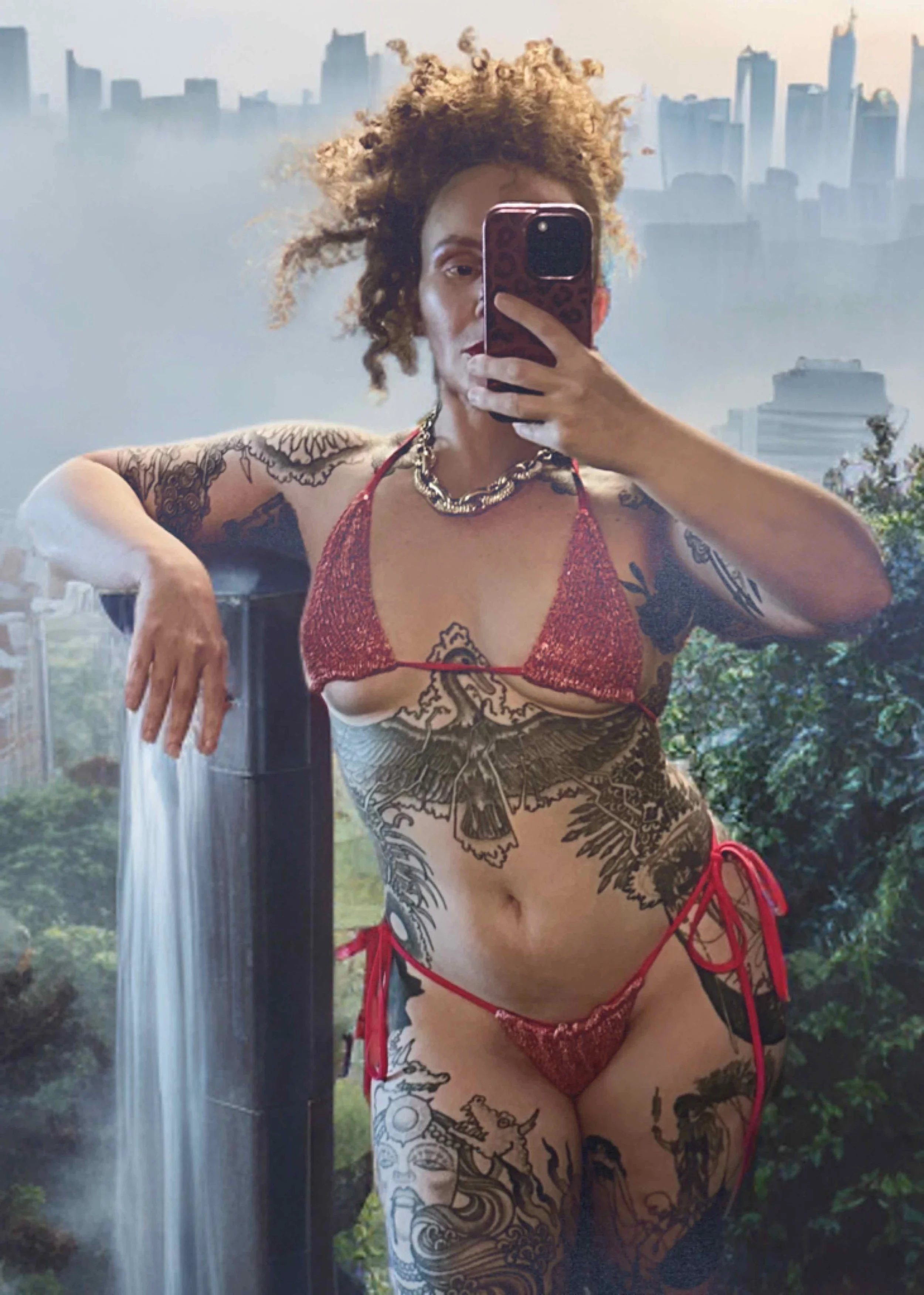 Alternative model with curly hair taking a mirror selfie in front of a city skyline backdrop, wearing a red bikini with tattoos on her arms, torso, and legs, holding a water feature.