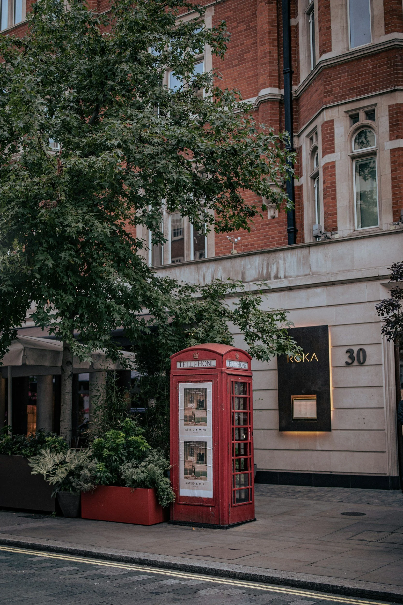 A red British telephone booth on a city sidewalk next to a building with a sign that reads 'ROKA 30' and leafy trees behind the booth.