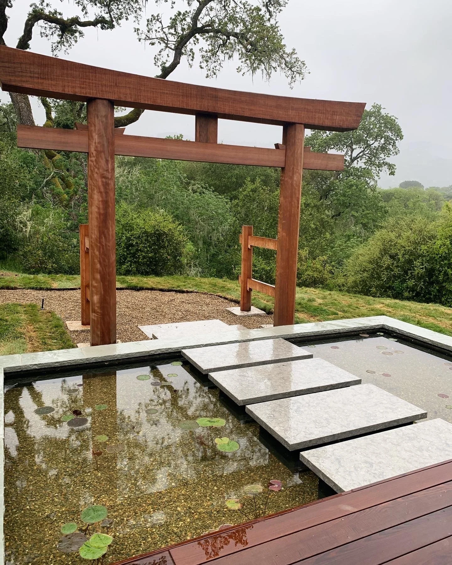 A traditional wooden Japanese torii gate in a natural outdoor setting with green trees and shrubs, near a pond with floating lily pads and stepping stones.