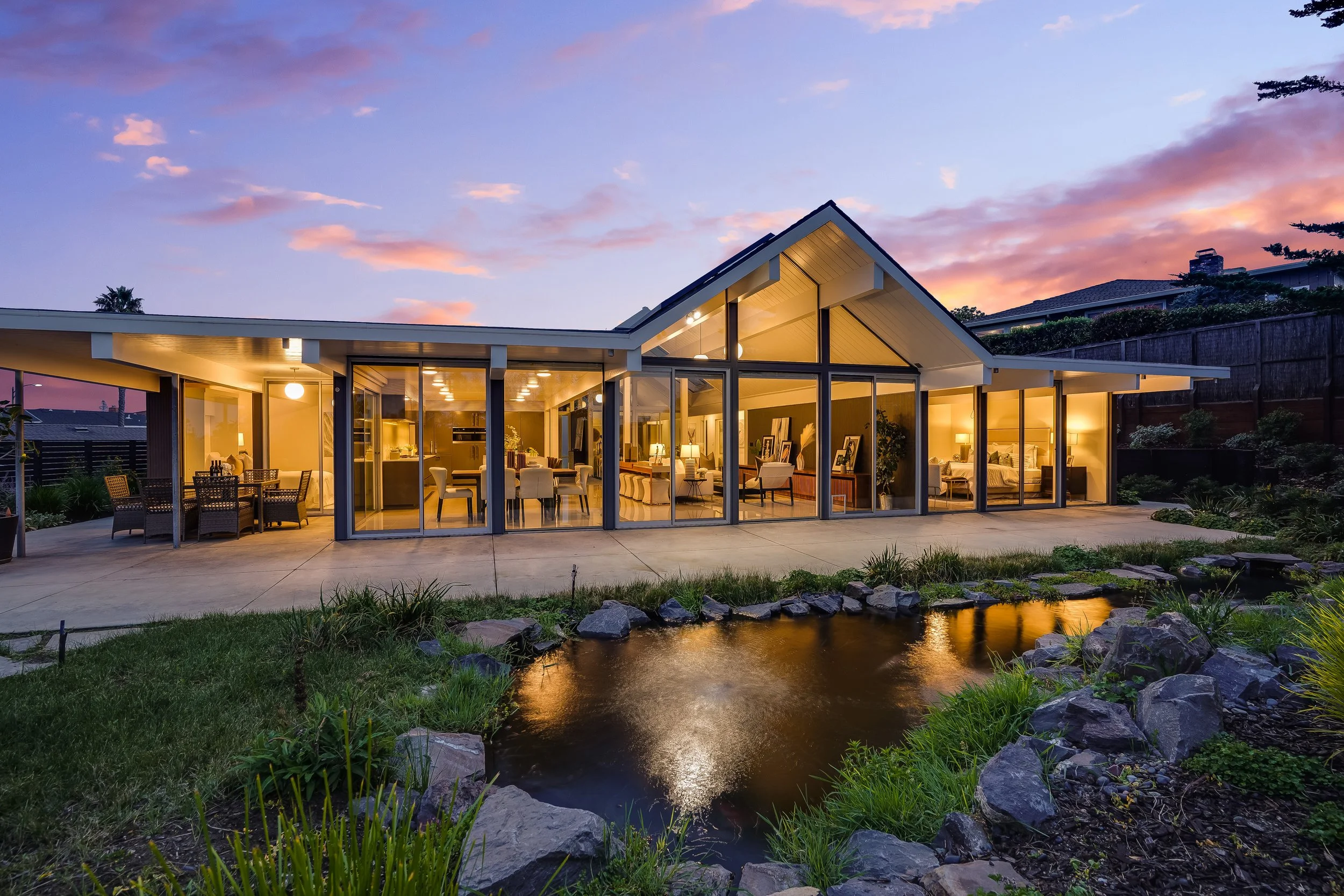 Modern house with large glass windows illuminated from inside, situated by a small pond with rocks and green plants, during sunset with colorful sky
