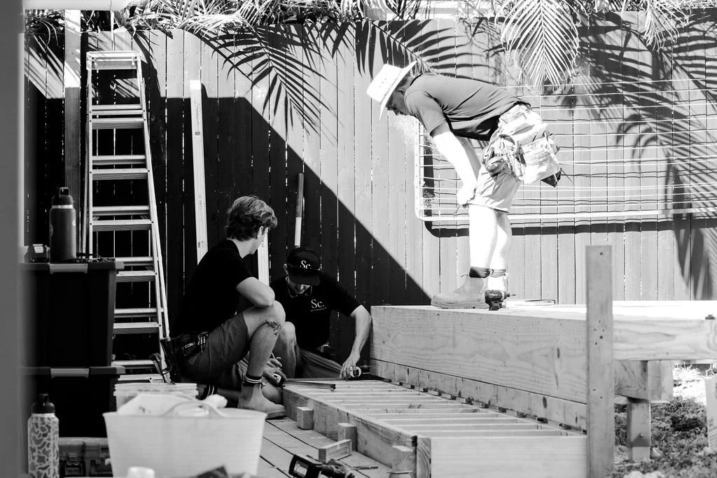 Three men working on building a deck, with one man on the elevated platform and two men kneeling on the ground, surrounded by construction tools and materials, with a wooden fence and tropical plants in the background.
