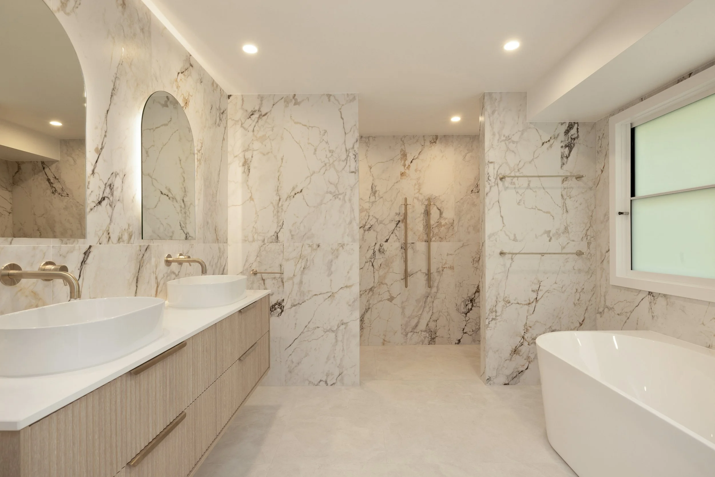 Modern bathroom with marble walls, dual vessel sinks on a wooden vanity, a large bathtub, a frosted window, and a walk-in shower with glass doors and metal handles.