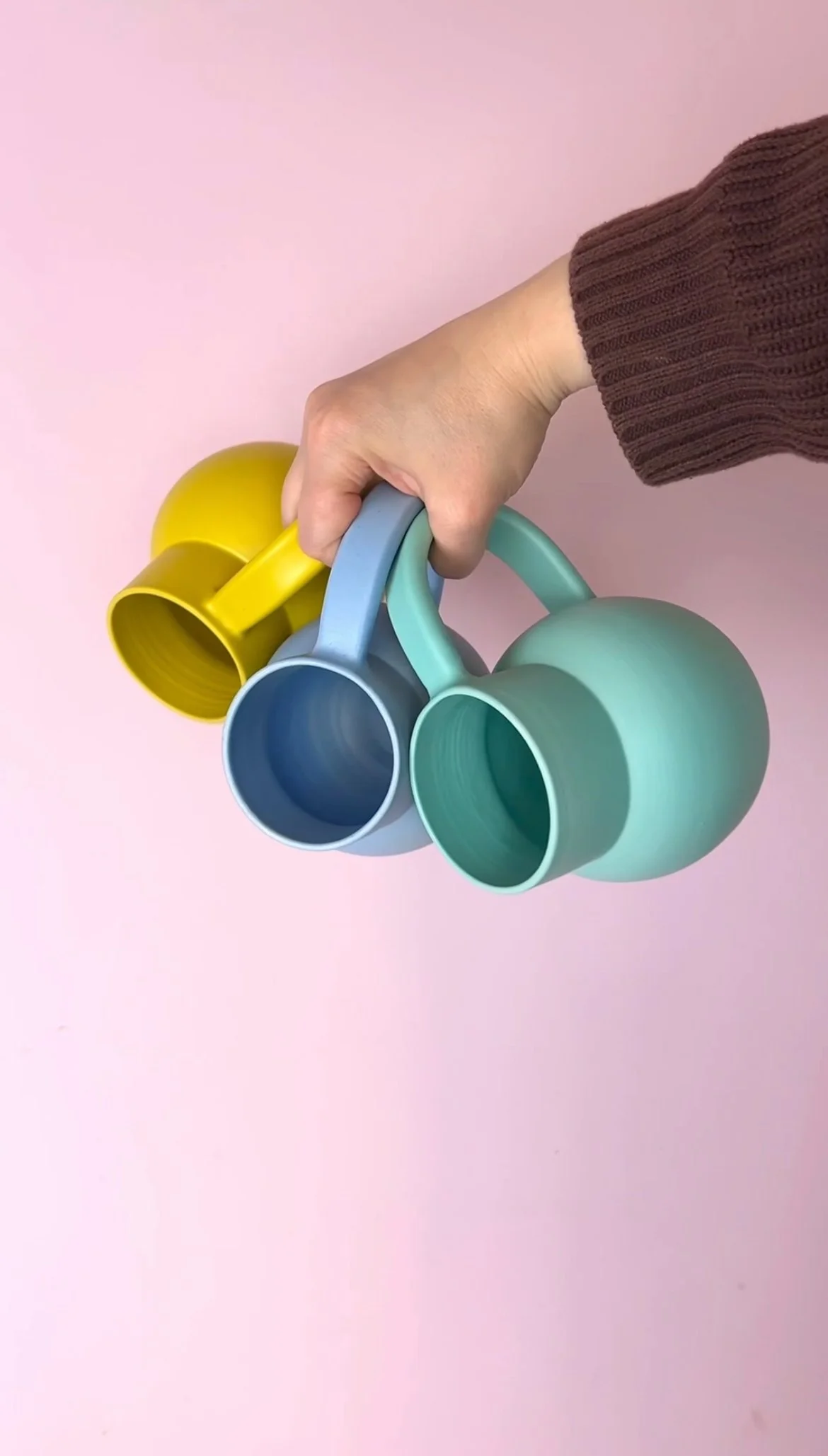 Marilyne Blais holding three colorful mugs in pastel shades of yellow, blue and teal against a pink background.