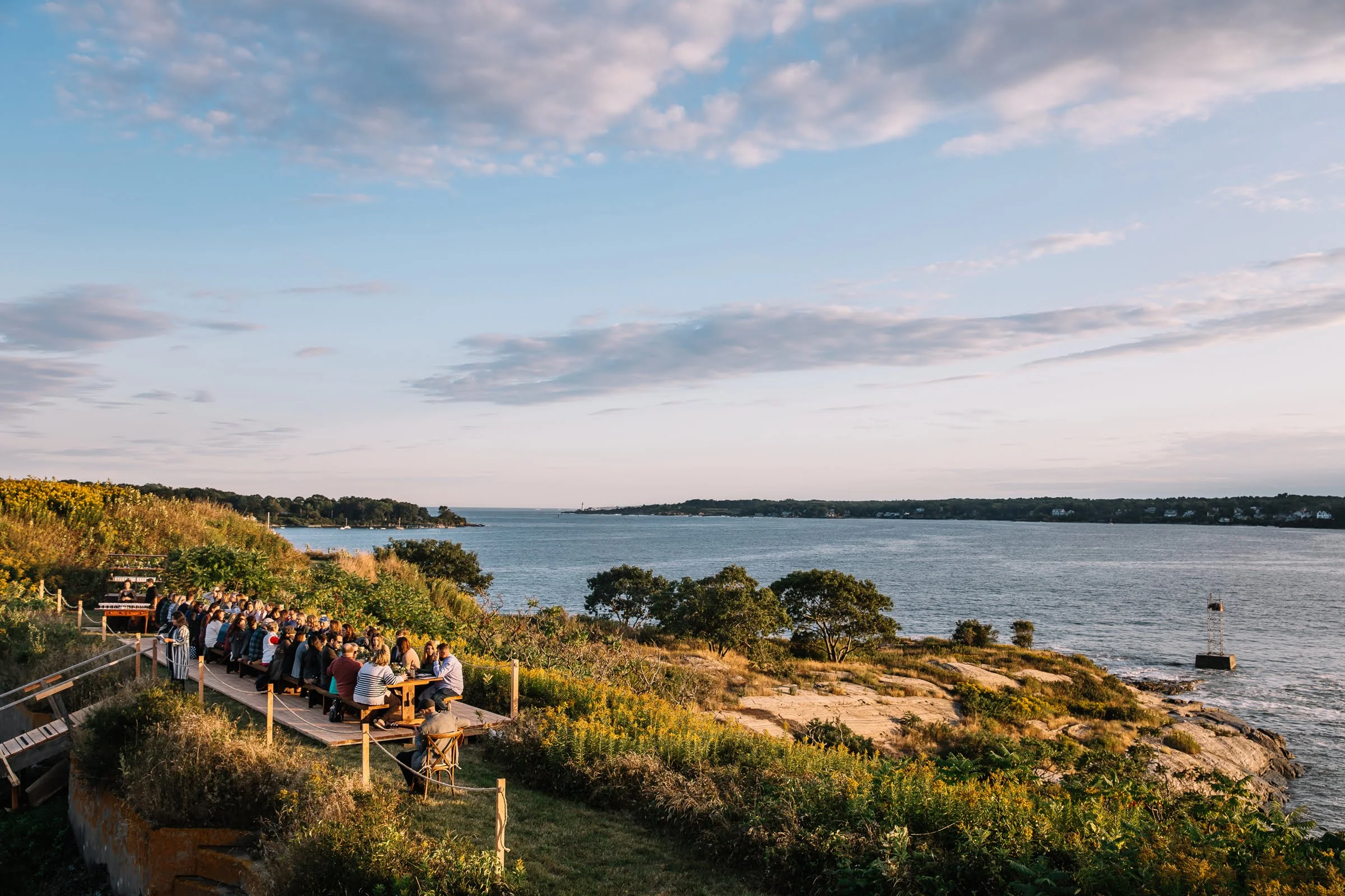A group of people sitting at long picnic tables on a wooden deck overlooking a body of water with green vegetation and a rocky shoreline, under a partly cloudy sky.