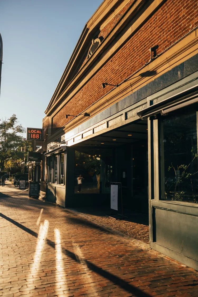 A brick building with large windows and a digital sign showing 'LOCAL 188' on a city street during sunset. The sidewalk and street are made of brick, with shadows cast by the building and nearby objects.