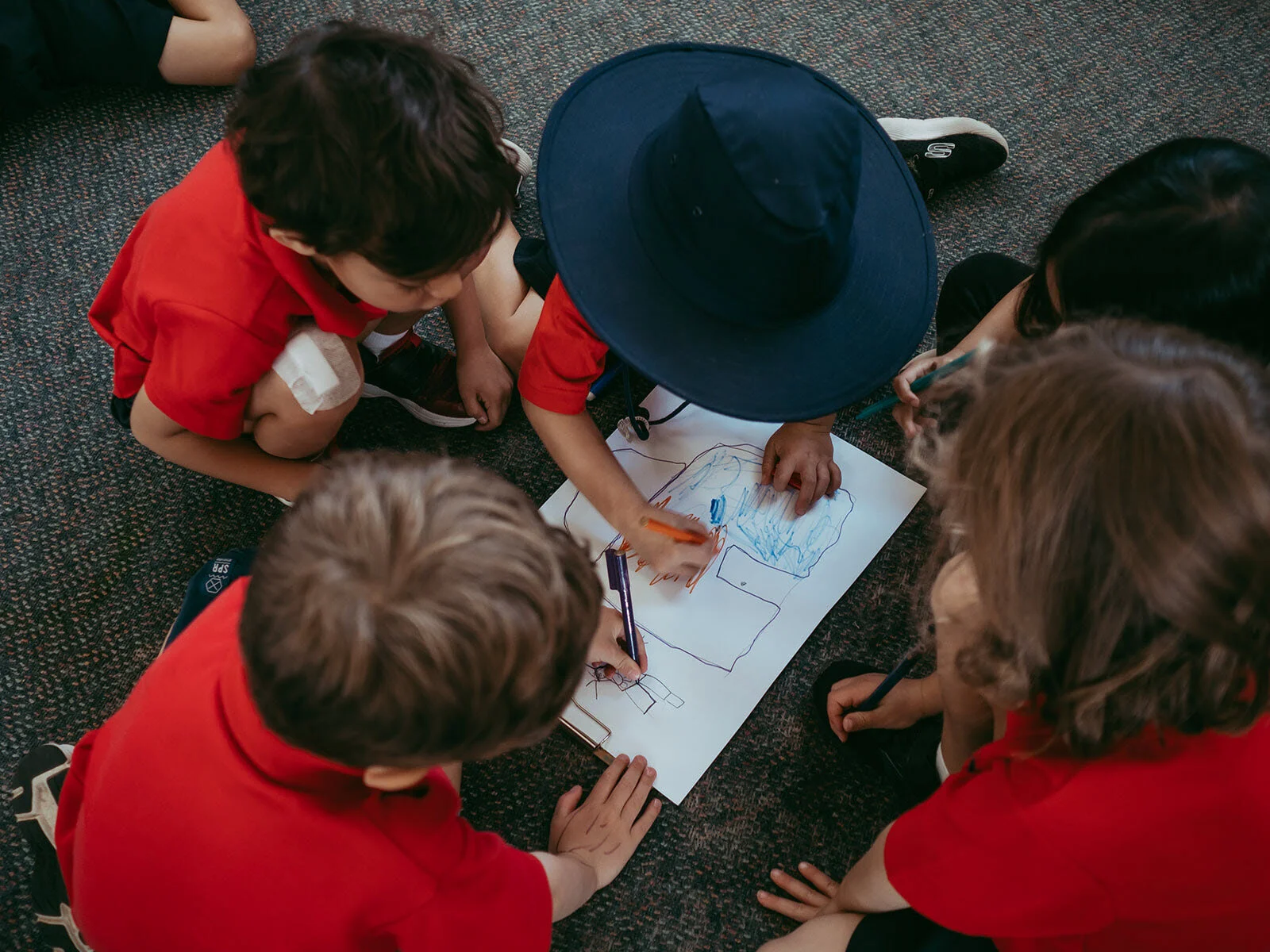 Group of children sitting on the carpeted floor, drawing on a large sheet of paper with colored markers.