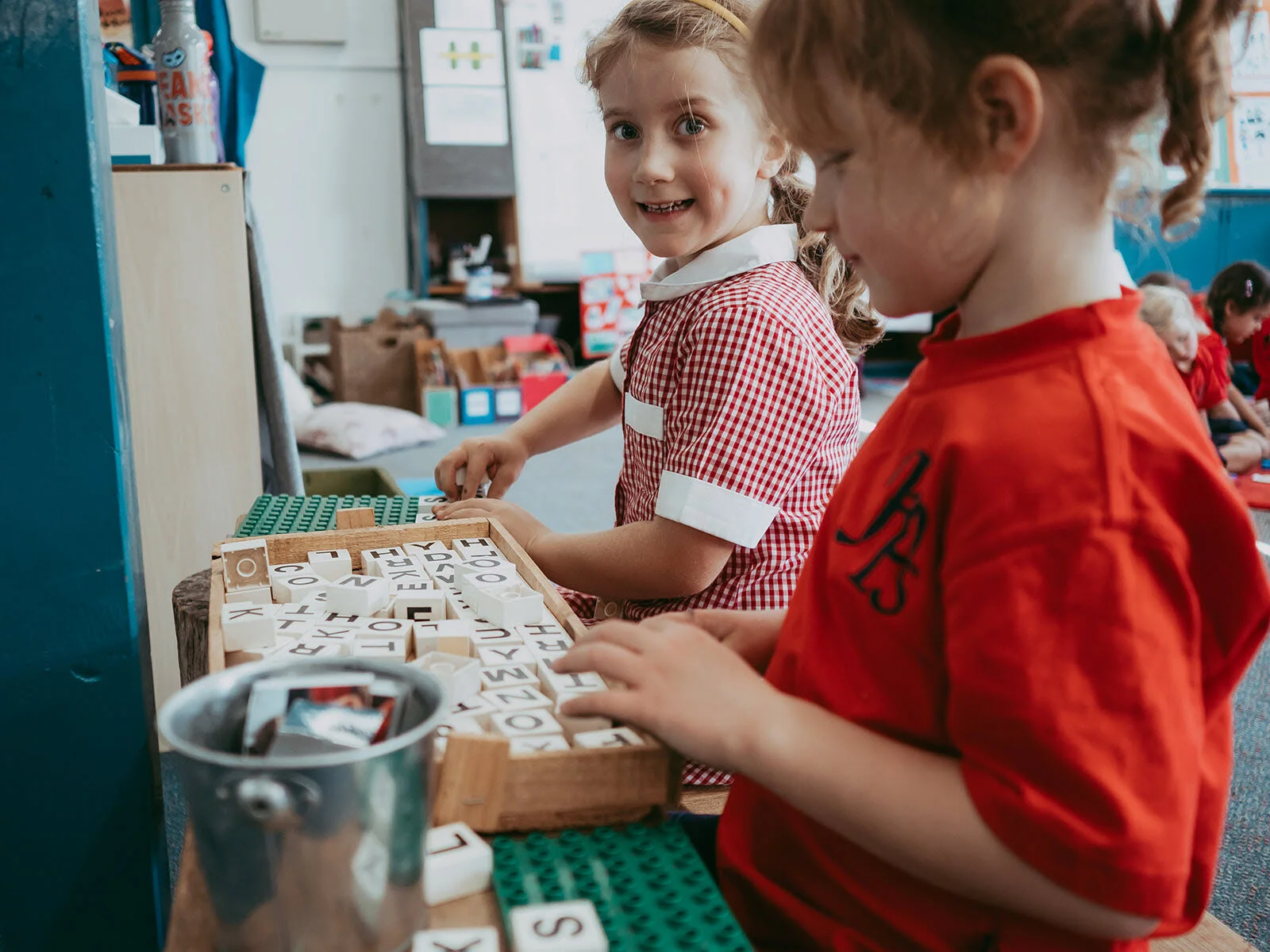 Two young girls playing with letter blocks at a table in a classroom, with other children in the background.