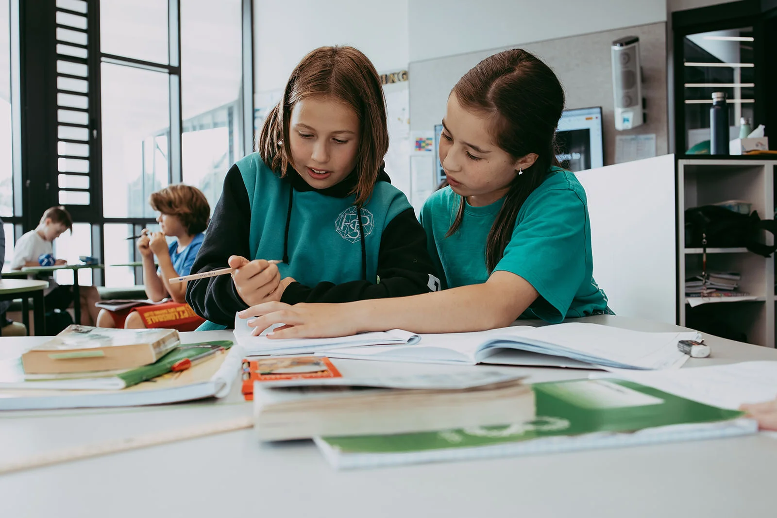 Two young girls studying together at a school desk with books and papers, in a modern classroom with large windows and other students in the background.